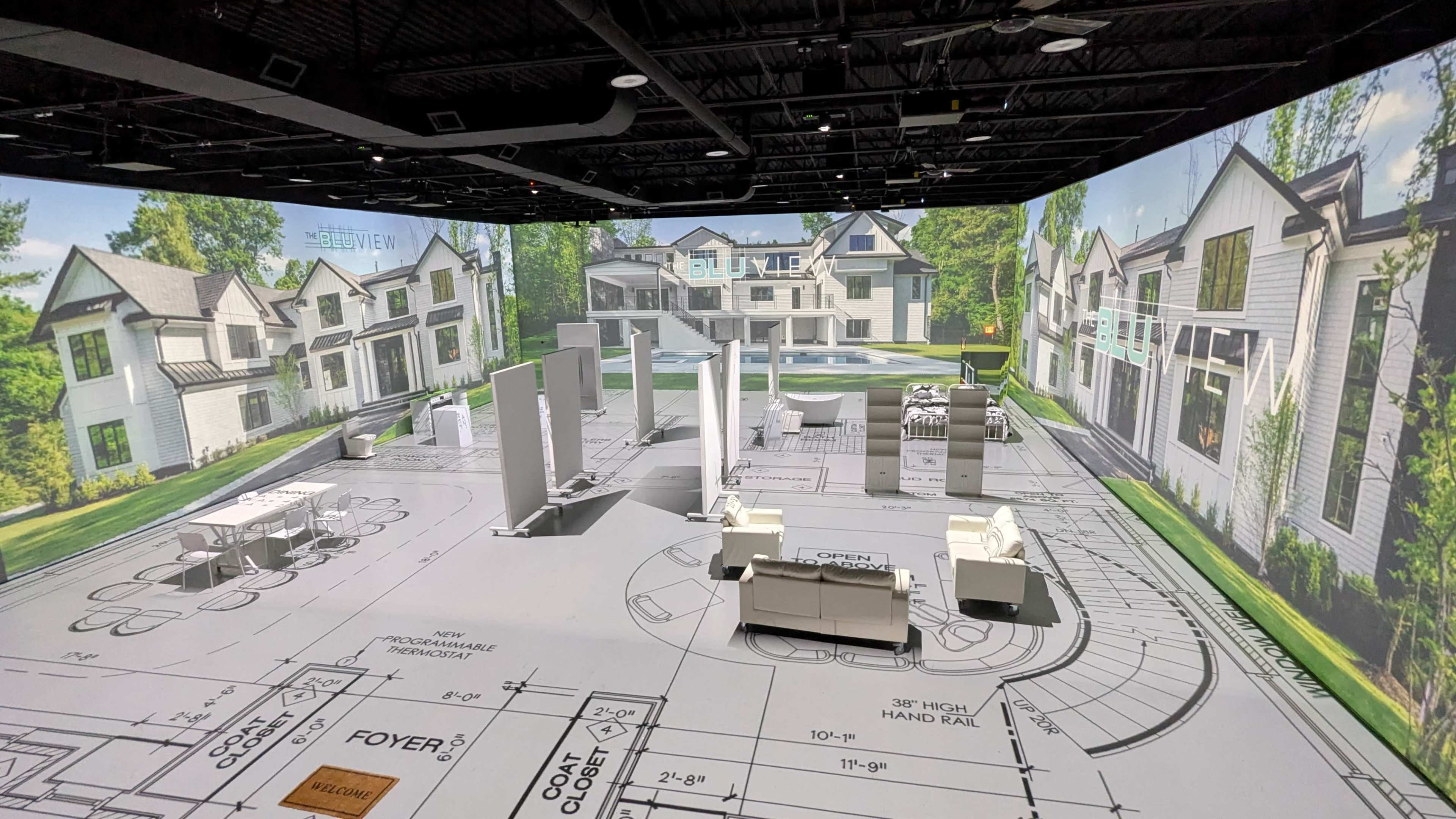 The image shows a large space designed as a virtual reality presentation area with architectural plans and rendered images of houses projected onto the walls.