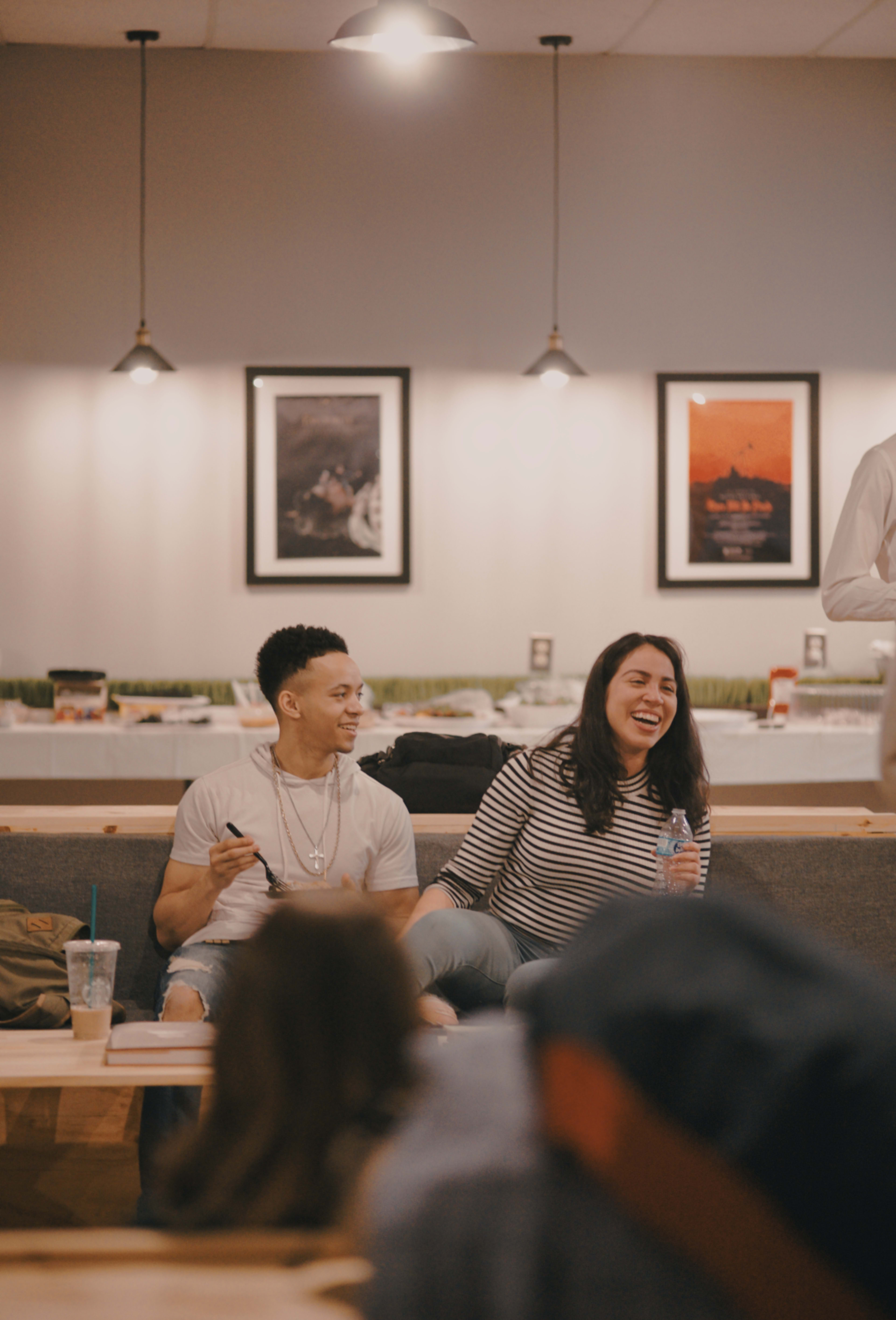 Two people are seated on a couch in a café, smiling and engaging with each other, while others are seen in the background.