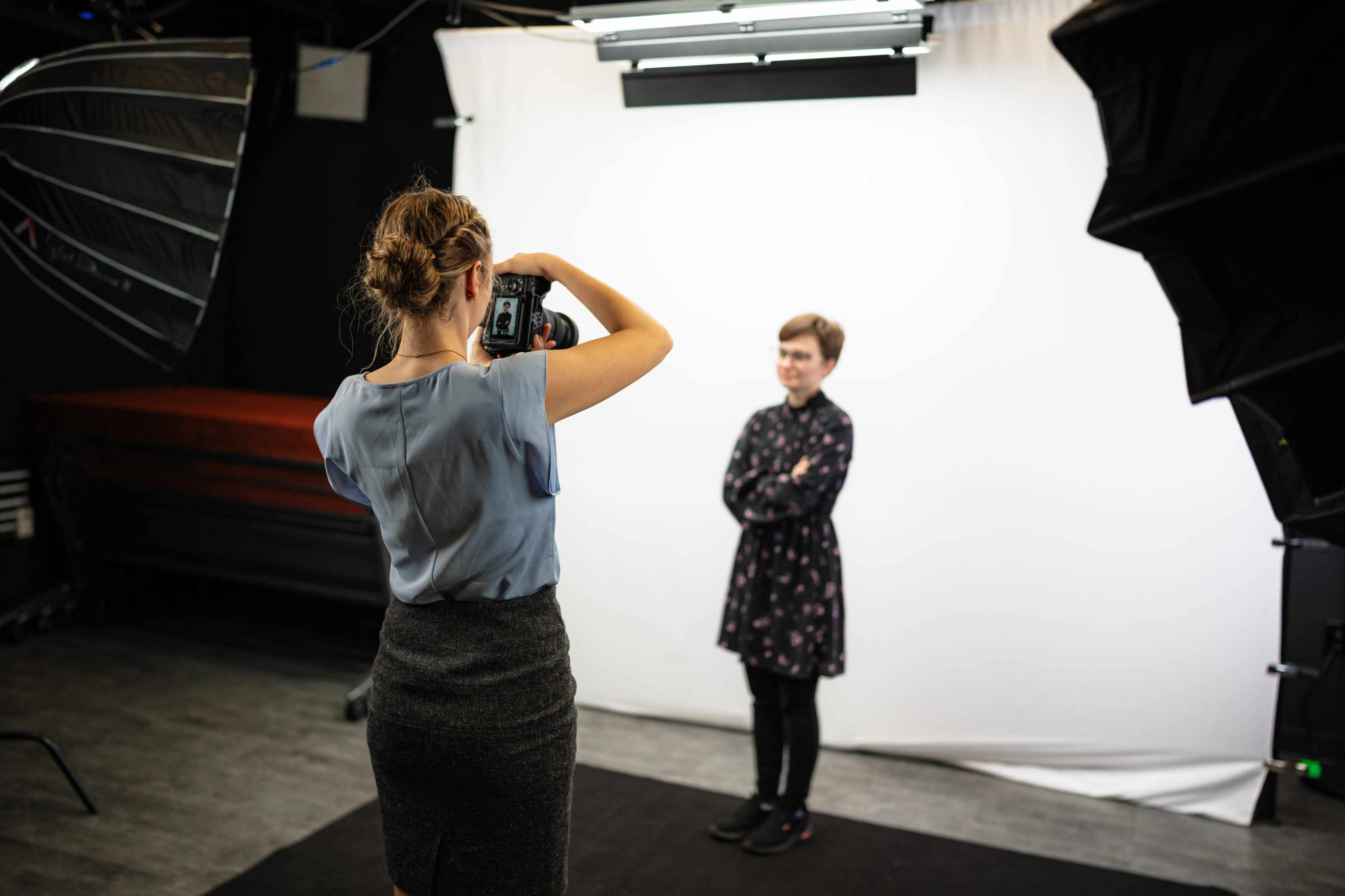 A photographer is taking a portrait of a person standing with crossed arms in front of a white backdrop in a studio.