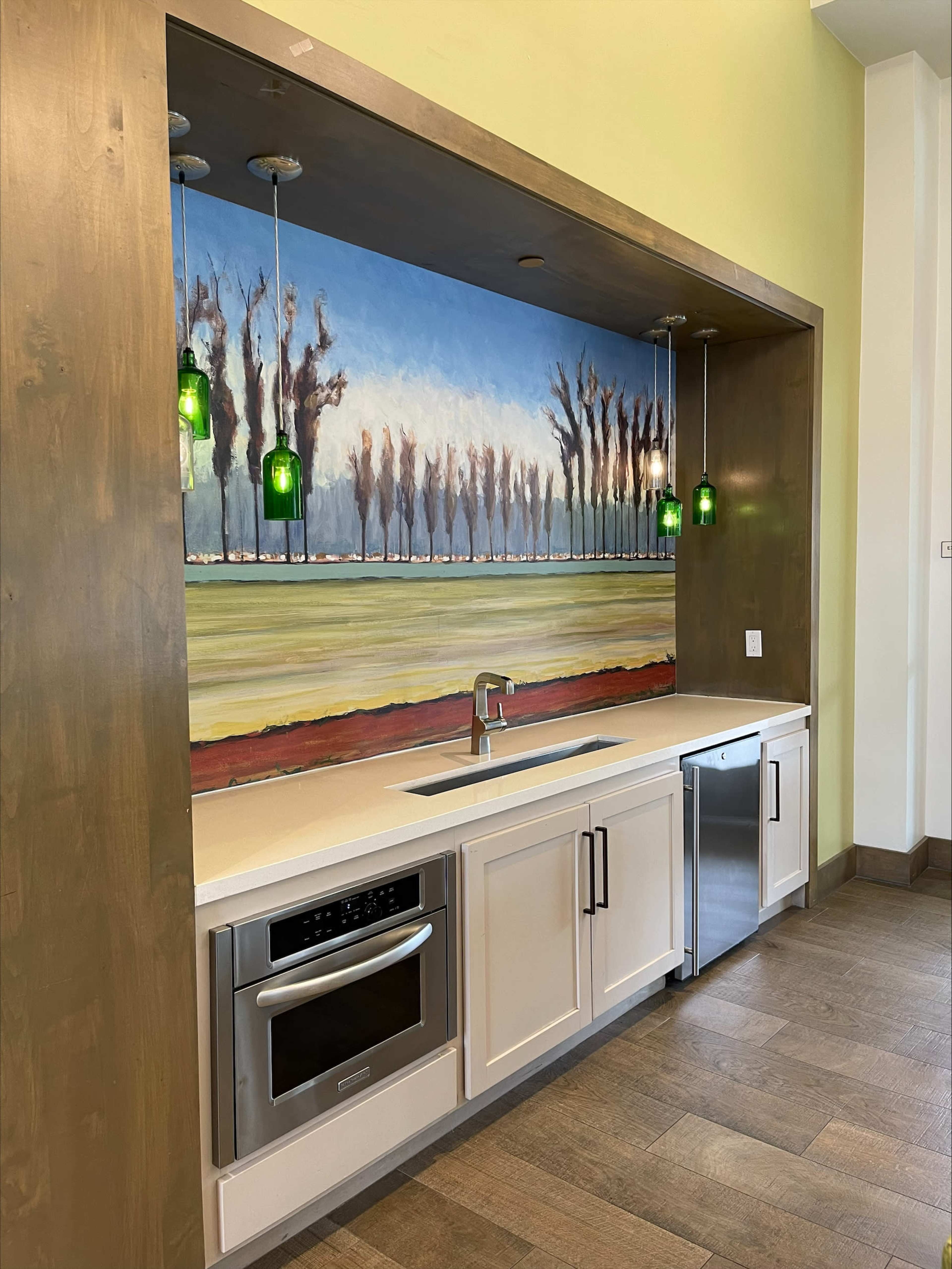 A kitchen area features a countertop with a sink, microwave, and dishwasher, set against a wall painted with a landscape and illuminated by green pendant lights.