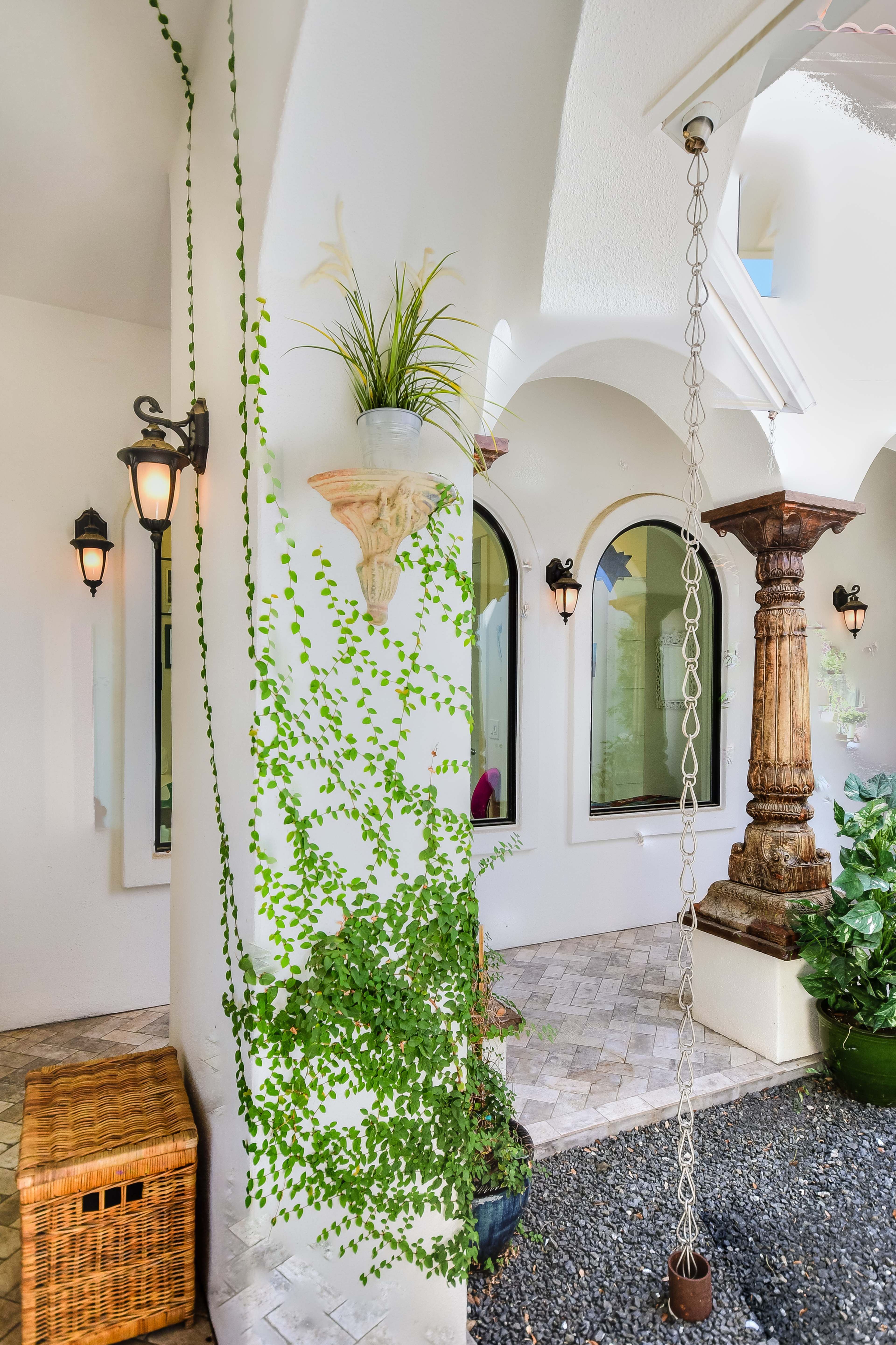 A courtyard with a decorative archway, flanked by green plants, a stone pillar, and lantern-style wall lights.