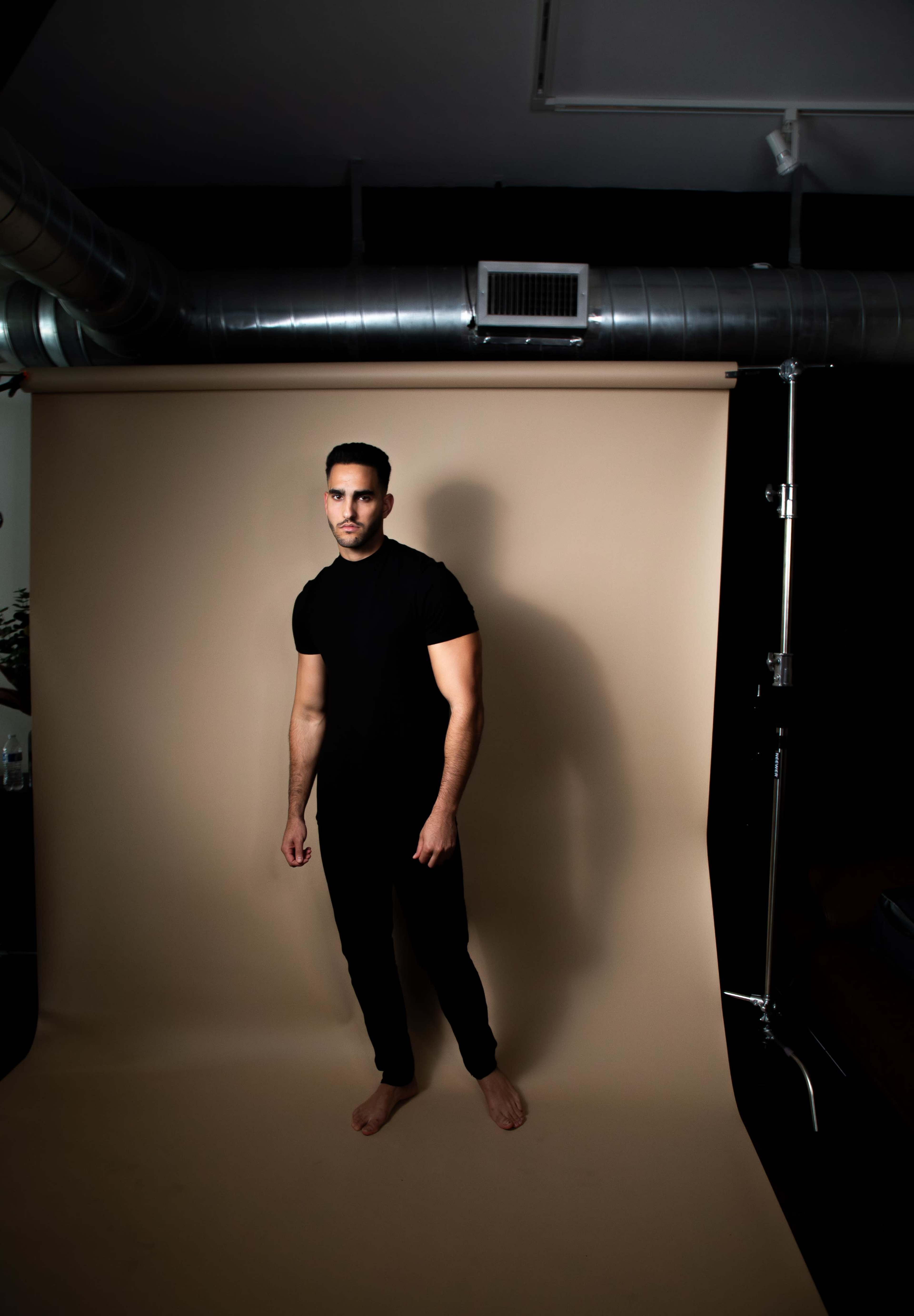 A man stands barefoot in a black room against a beige backdrop.