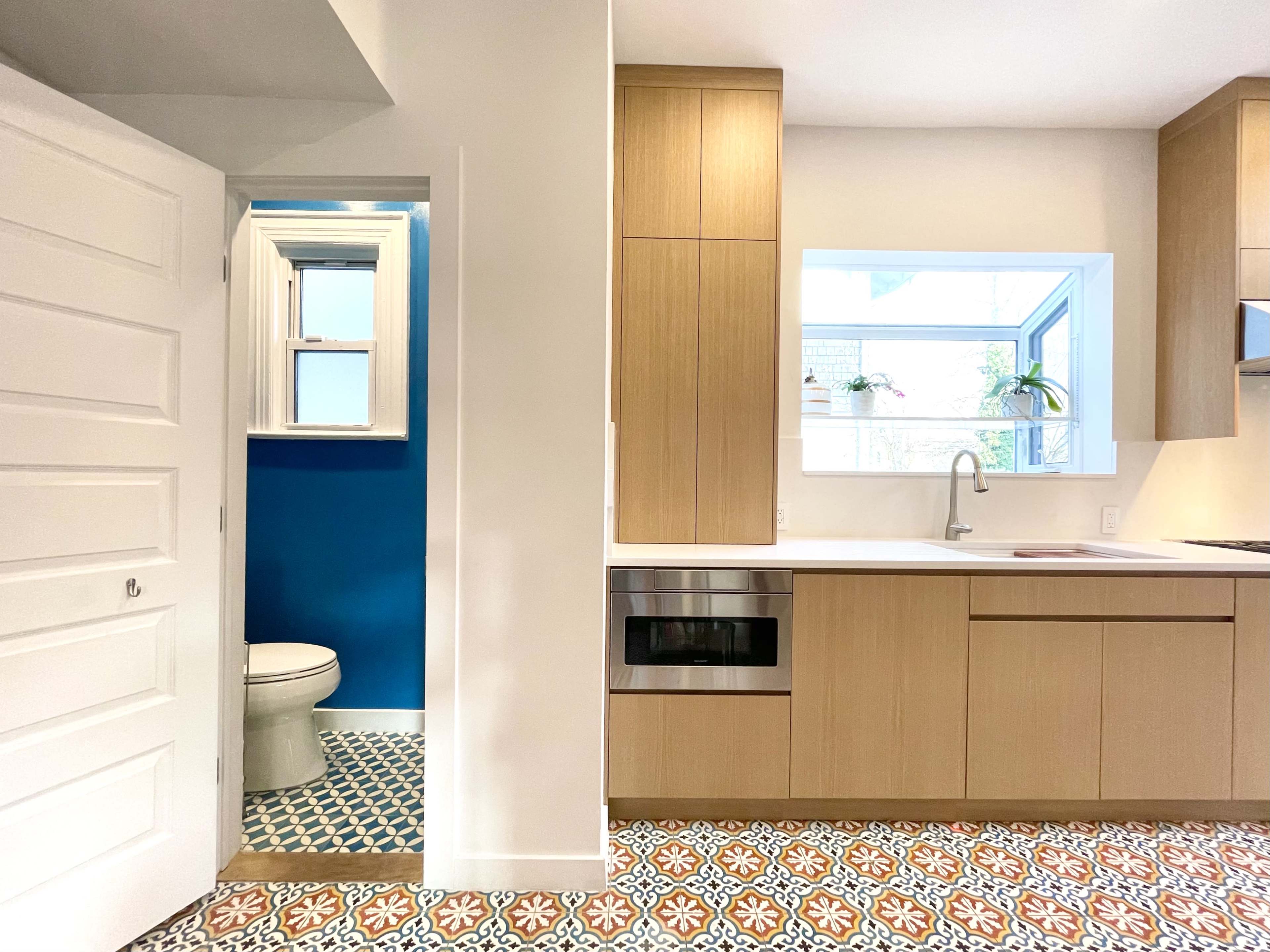 A modern kitchen features wooden cabinetry, a white countertop, and a view of a blue-painted bathroom through an open door.
