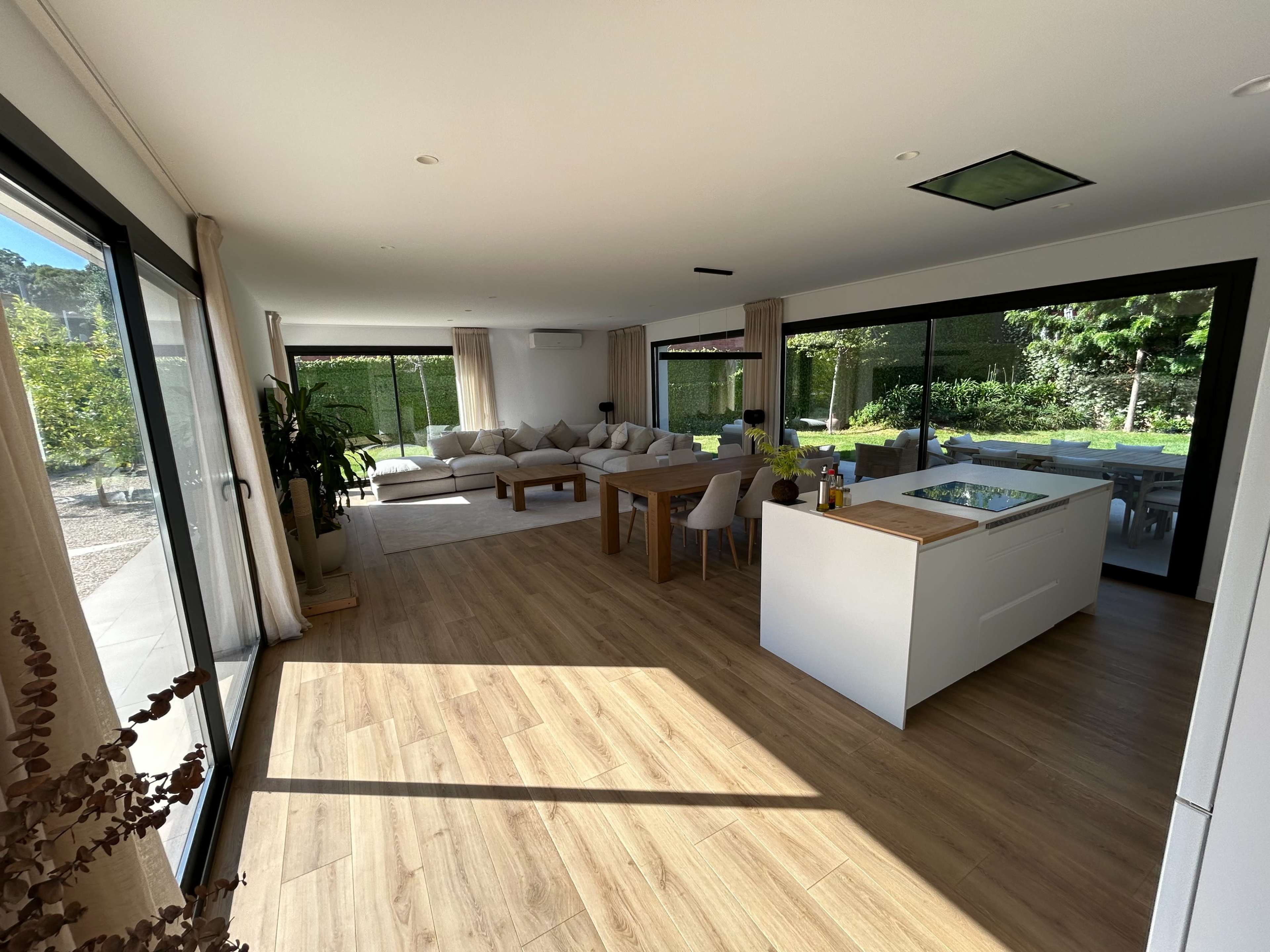 The image shows a modern, open-concept living space with a white kitchen island, a dining table, and a seating area that overlooks a garden through large glass windows.