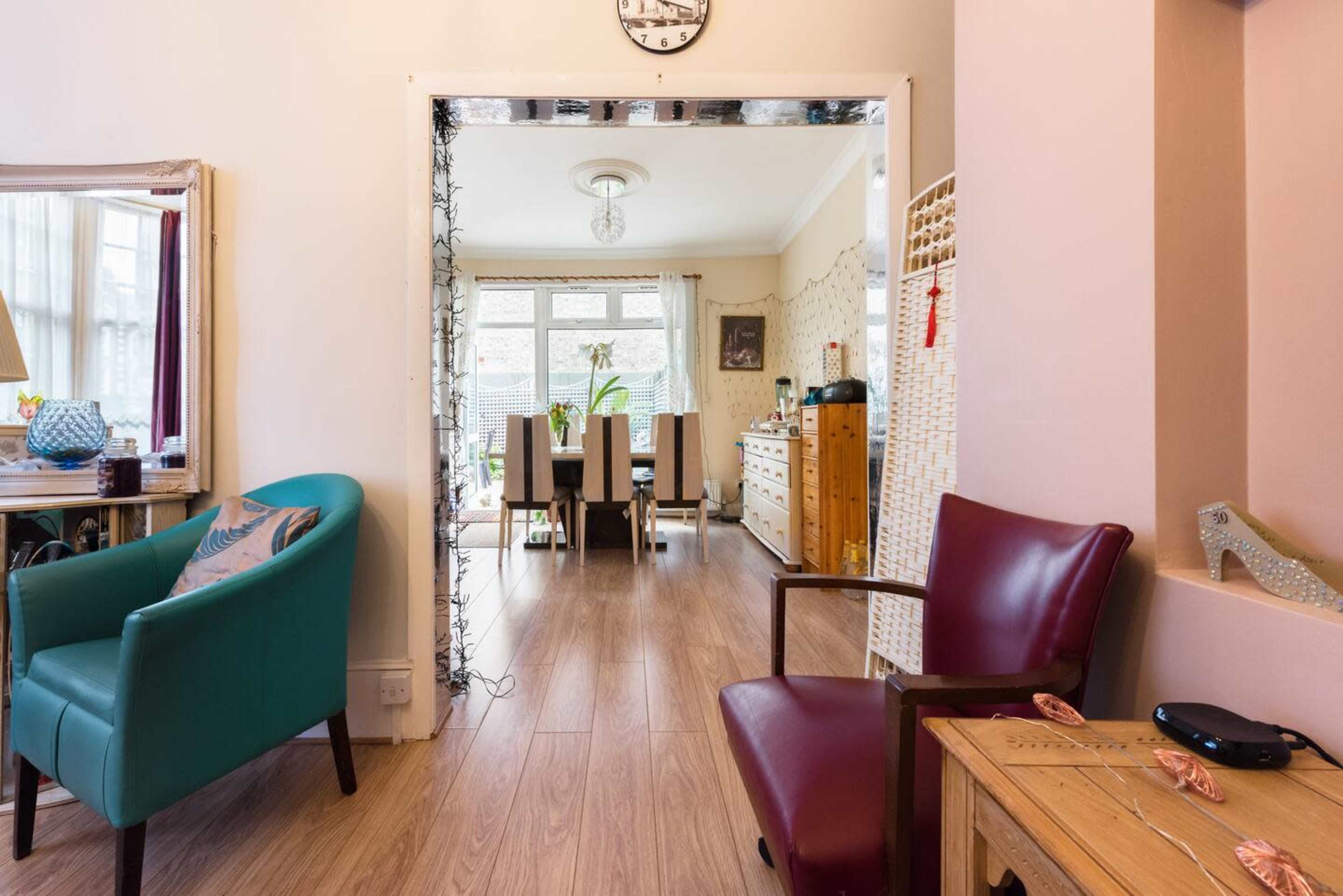 The image shows a cozy living space with two chairs, a wooden table, and a view of a bright dining area in the background.