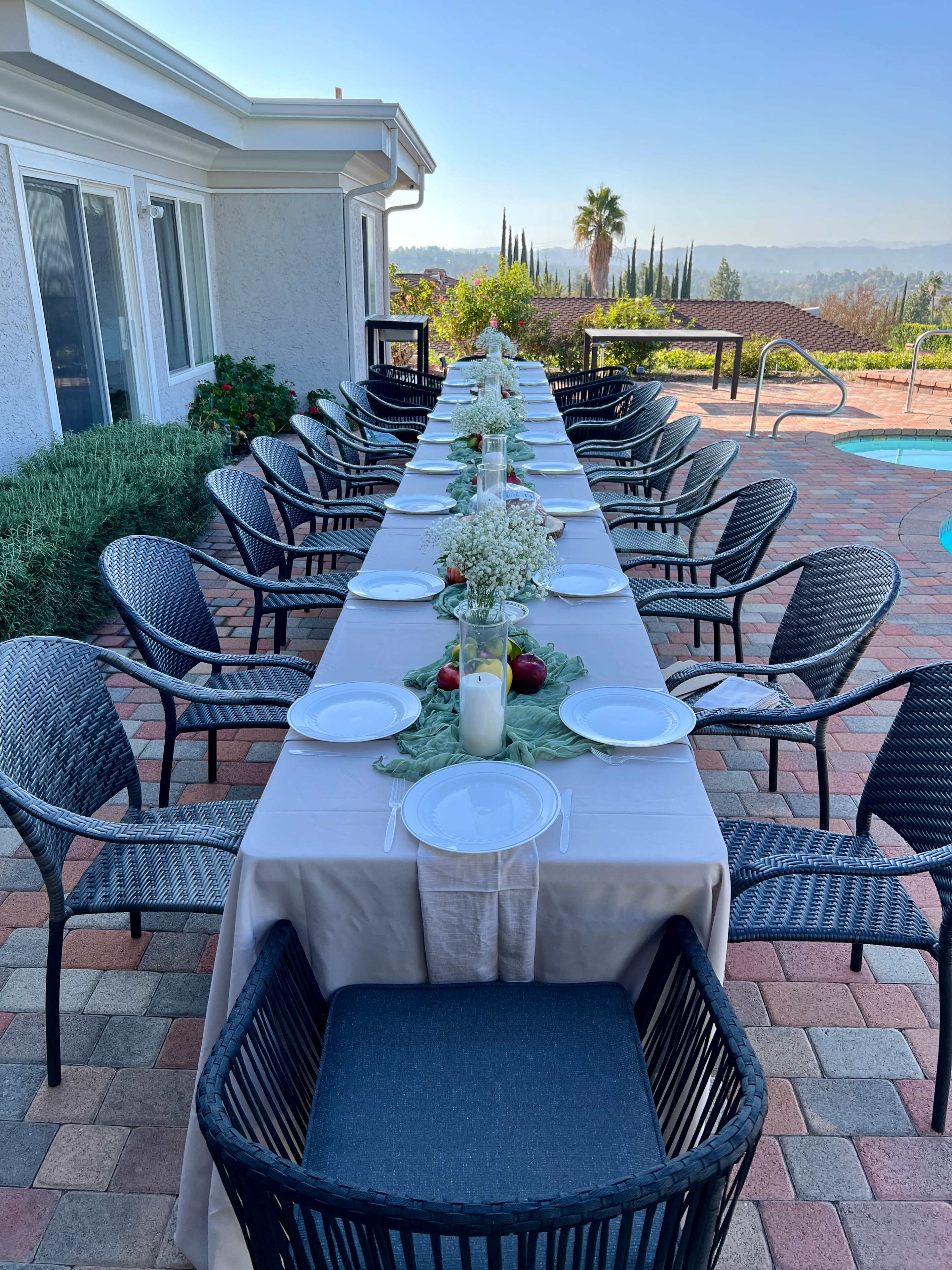 A long dining table is set outdoors beside a pool, with empty chairs and plates arranged along the table.