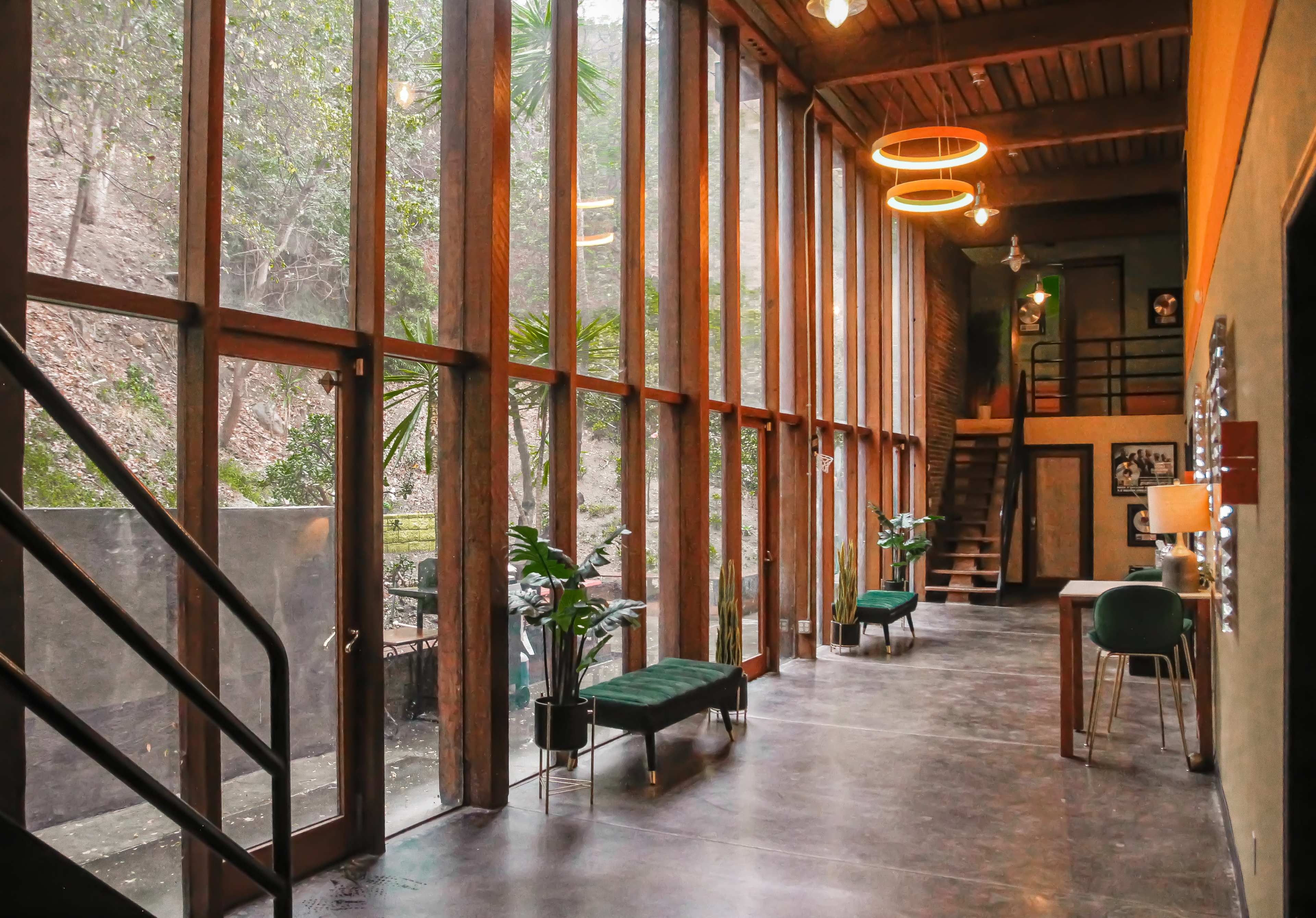 A spacious corridor with large windows, staircases, and minimalistic furniture is lined with plants and illuminated by circular light fixtures.