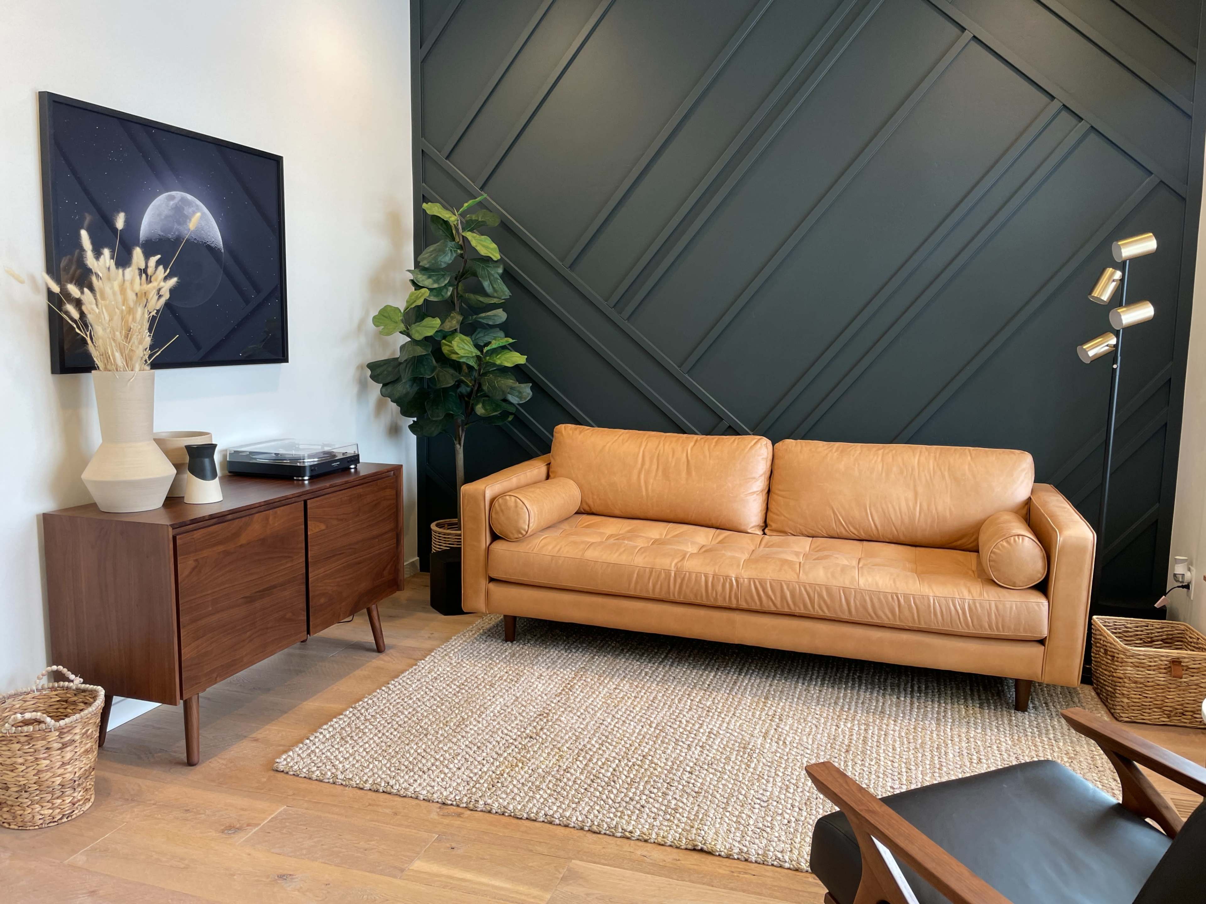 The scene features a modern living room with a tan leather sofa, a wooden sideboard, and decorative plants against a dark green accent wall.