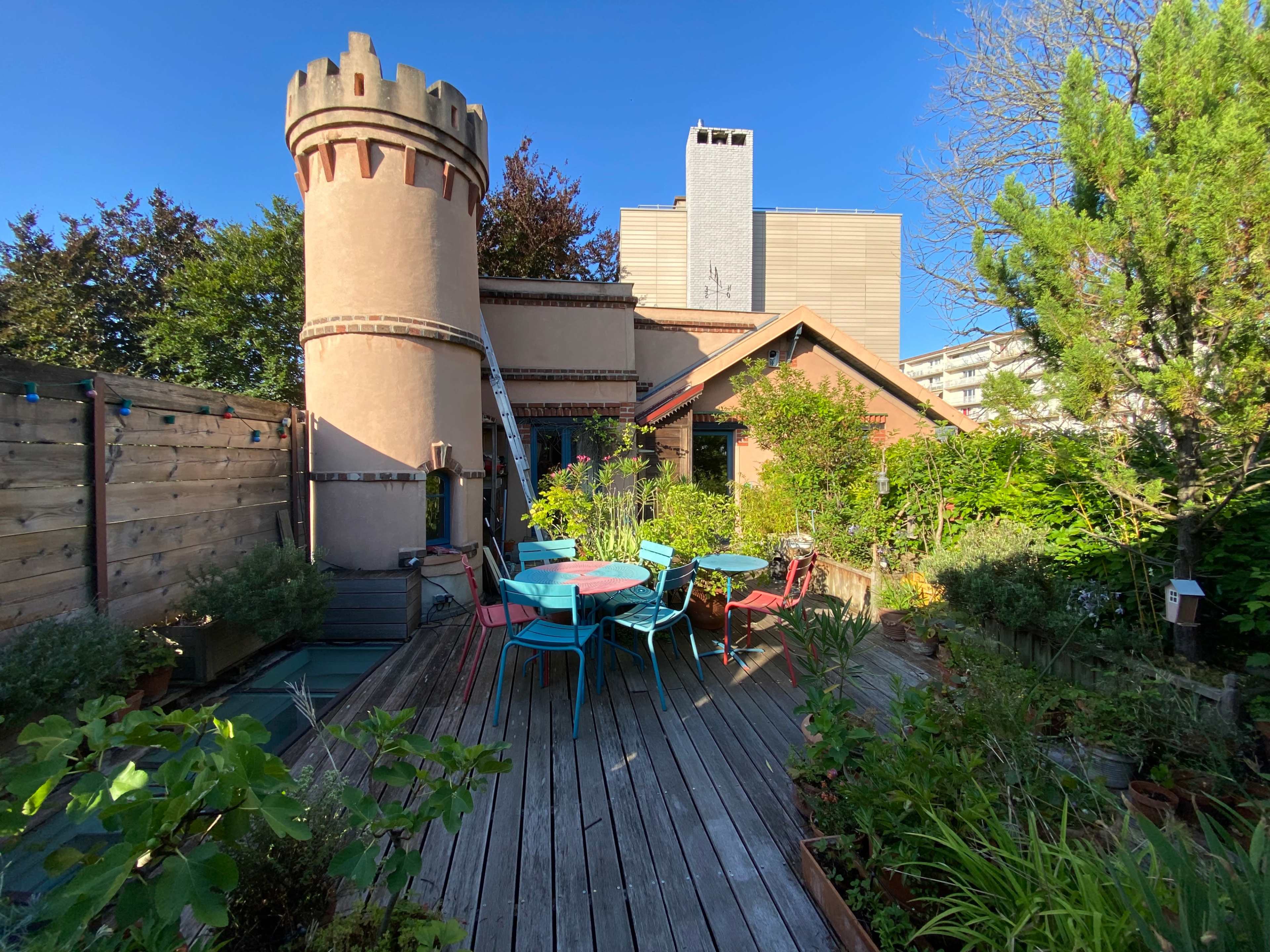 The image shows a wooden deck garden with colorful chairs and tables, surrounded by various plants and a tower-like structure in the background.
