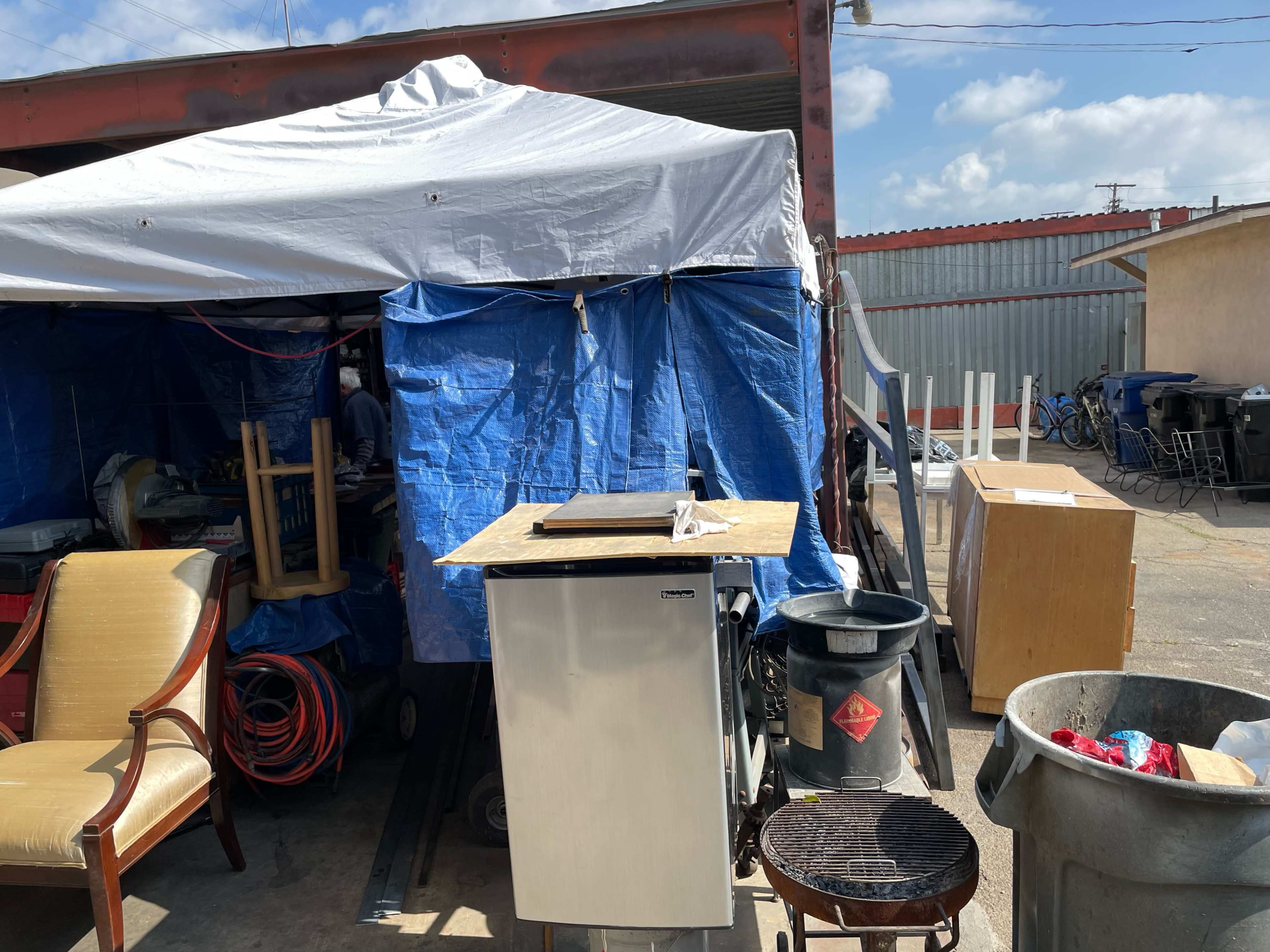 The image shows a large tent structure with blue tarps and various items, including a small refrigerator and a grill, situated in a cluttered outdoor area.