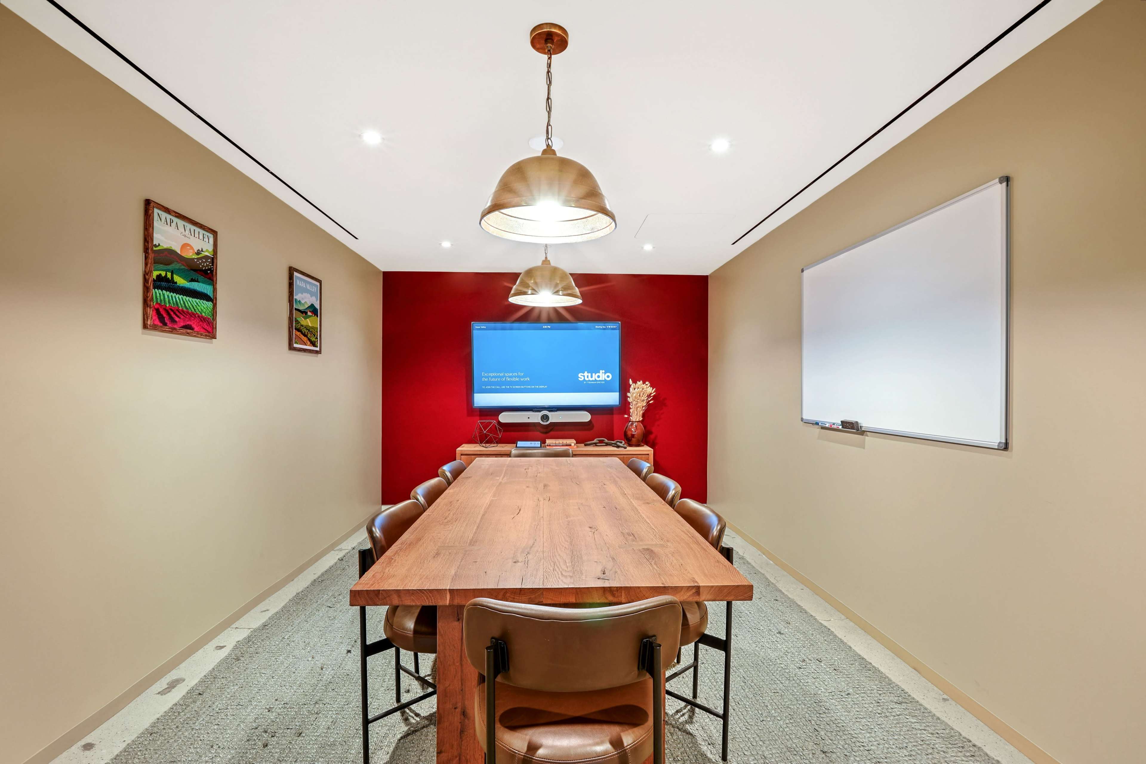 A long wooden conference table is surrounded by brown chairs in a small meeting room featuring a large screen on the wall and a whiteboard.
