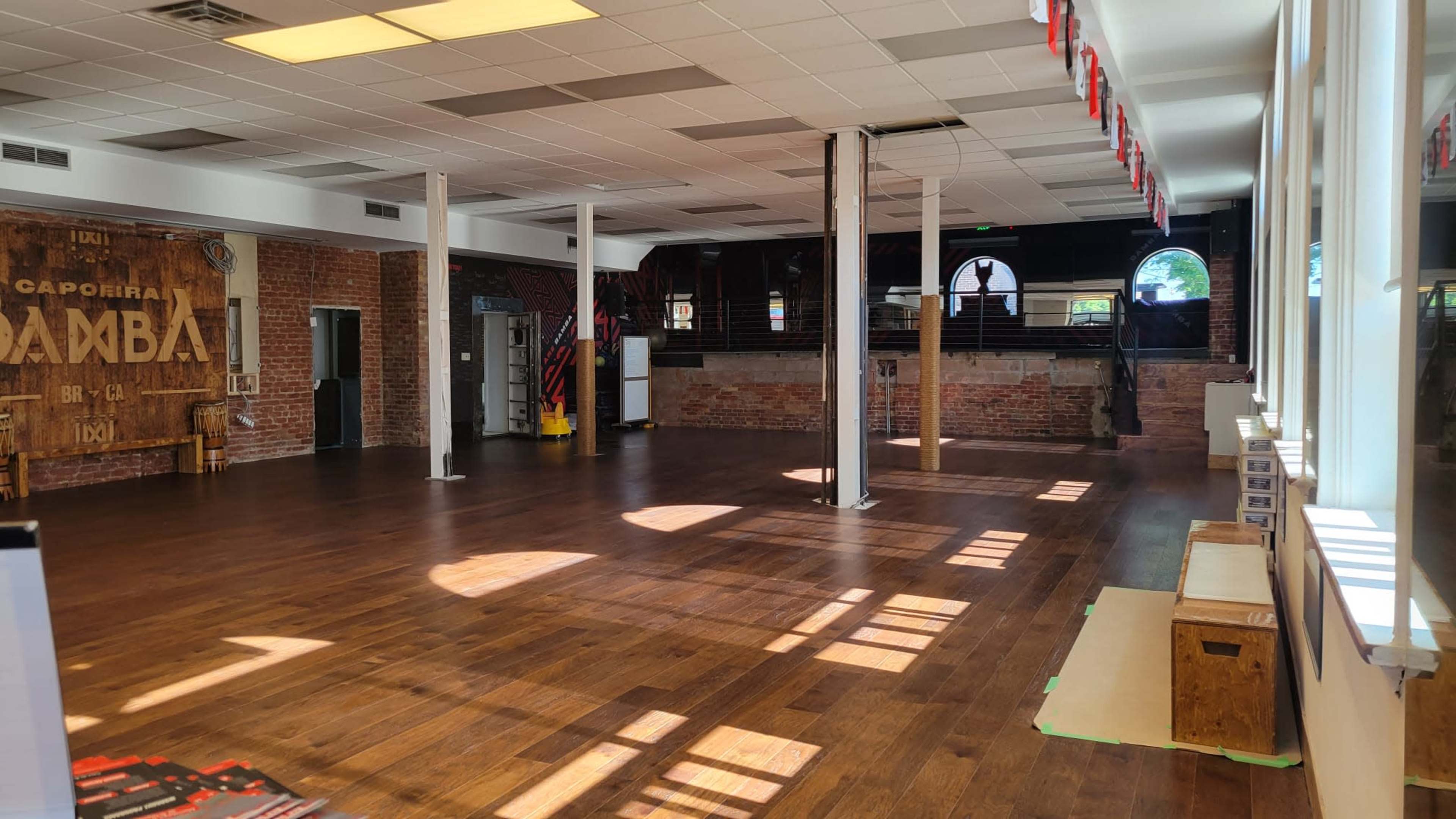 A spacious dance studio with wooden floors, bright natural light streaming in through large windows, and exposed brick walls.