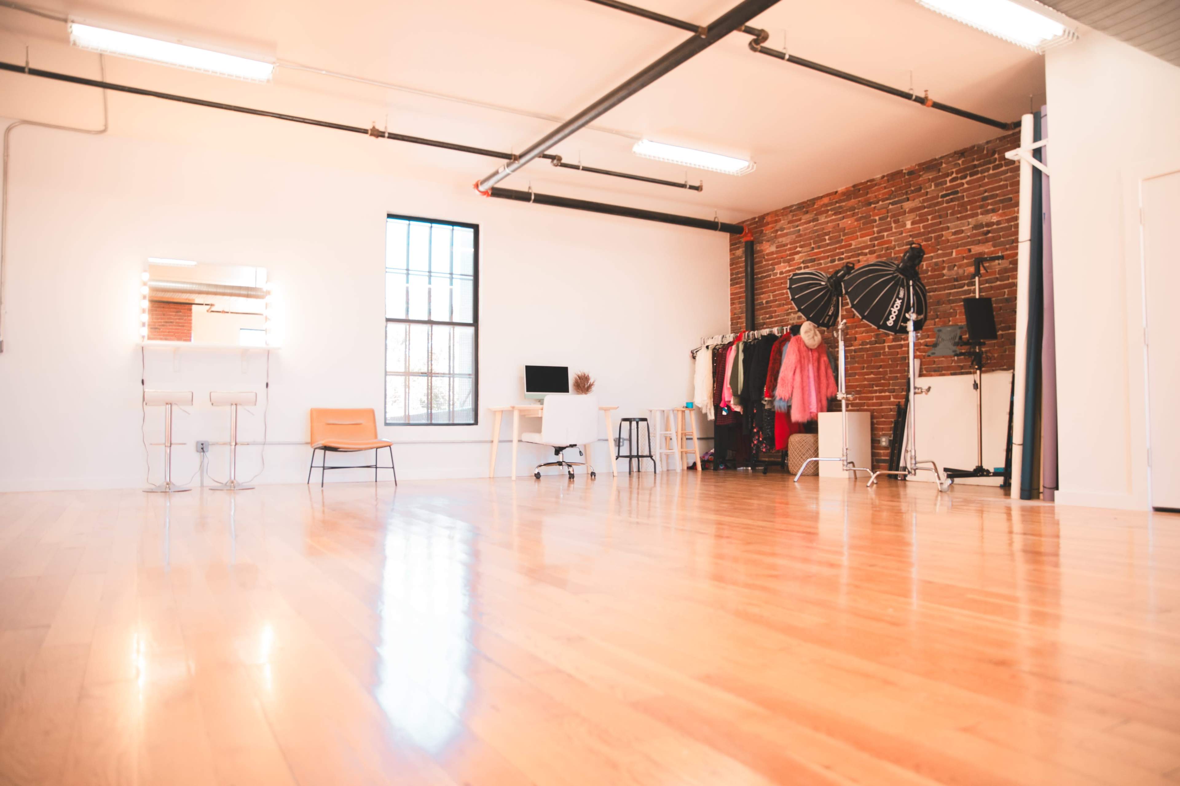 The image shows a spacious studio with hardwood floors, a brick wall, and minimal furniture, including a desk, chairs, and a clothing rack.
