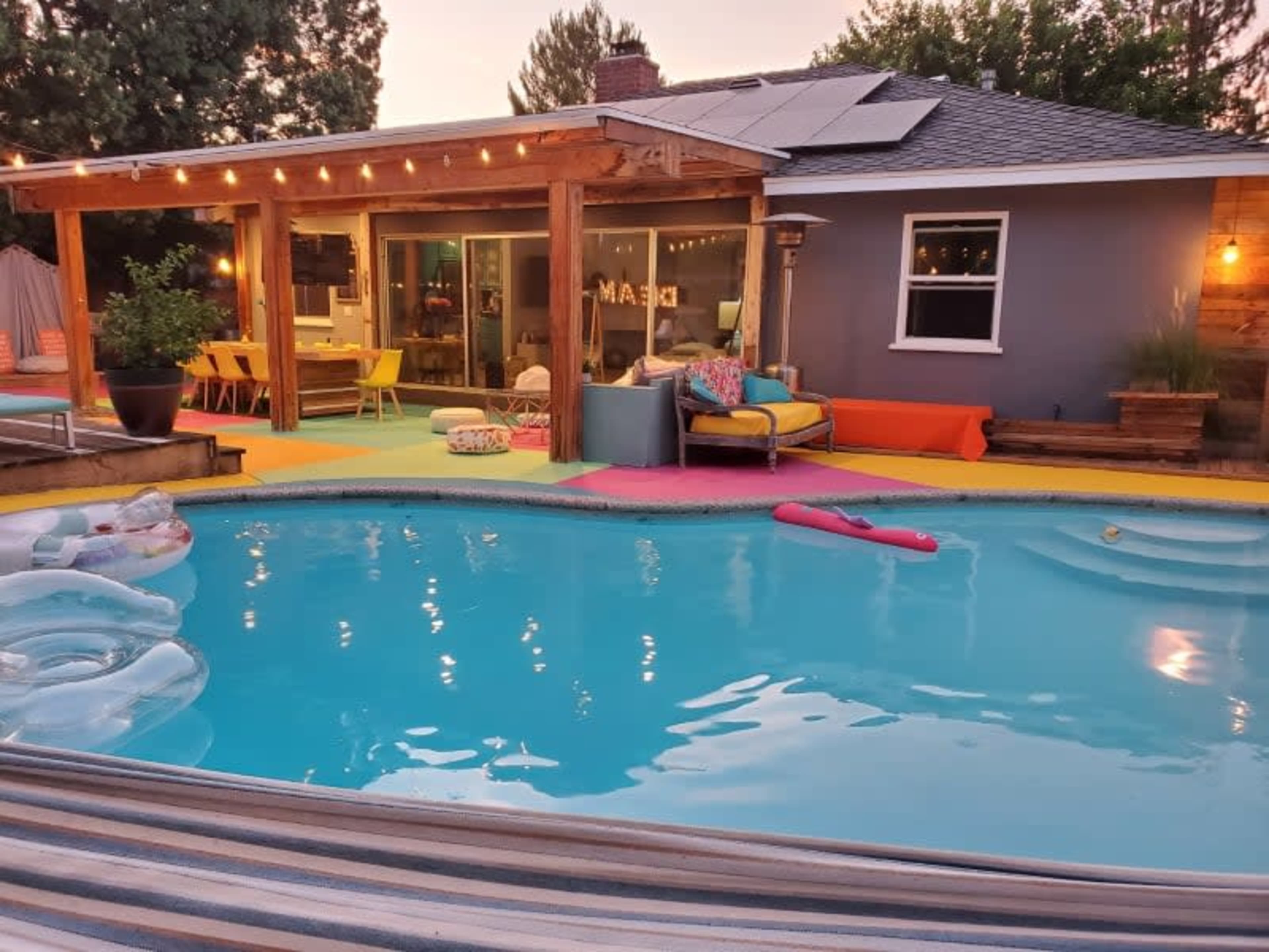 The image shows a backyard with a swimming pool, colorful loungers, and a house featuring large windows and a patio under string lights.