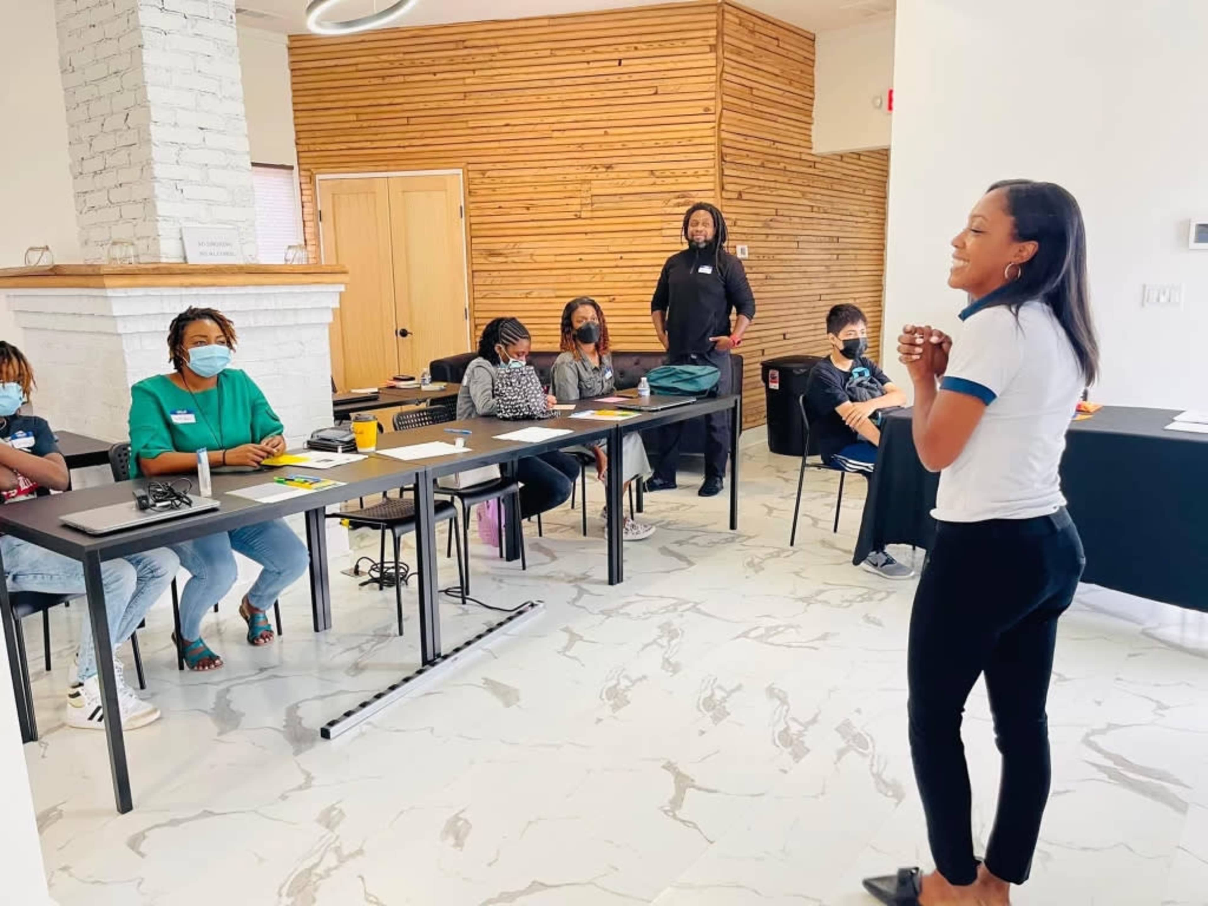 A woman stands in front of a group seated at tables in a classroom setting, while a man is standing in the background.