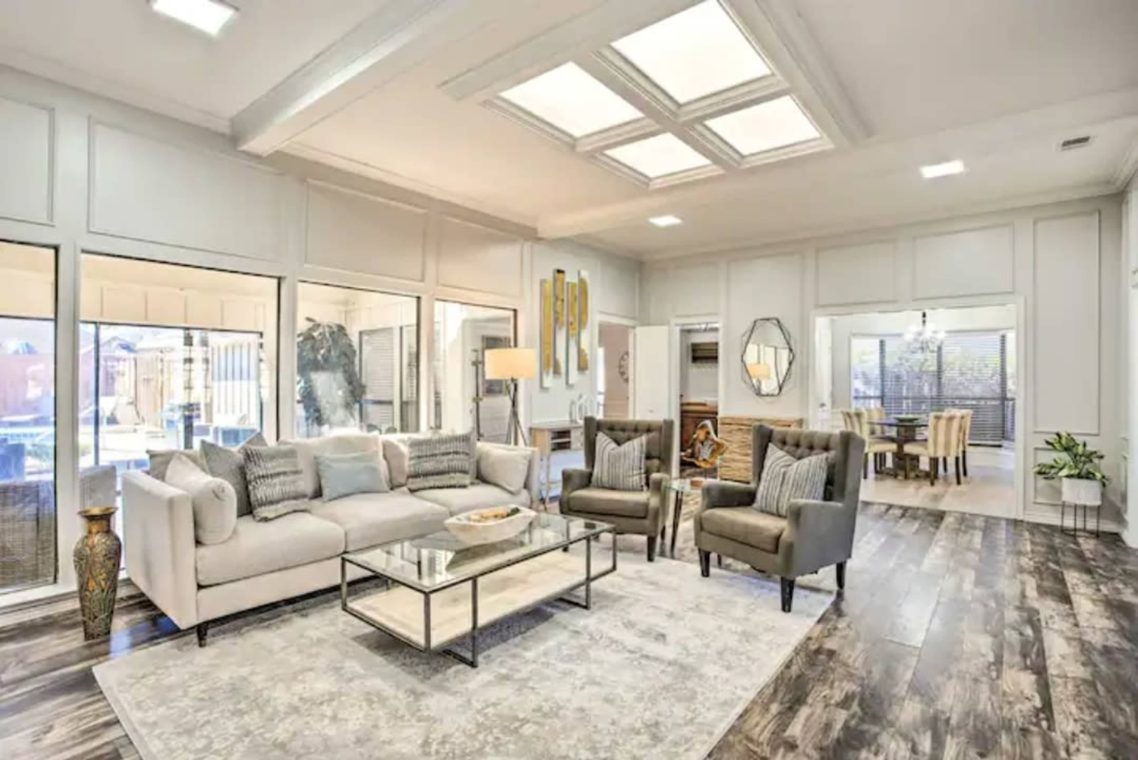 The living room features a light-colored sofa, two armchairs, a glass coffee table, and large windows that overlook a patio area.