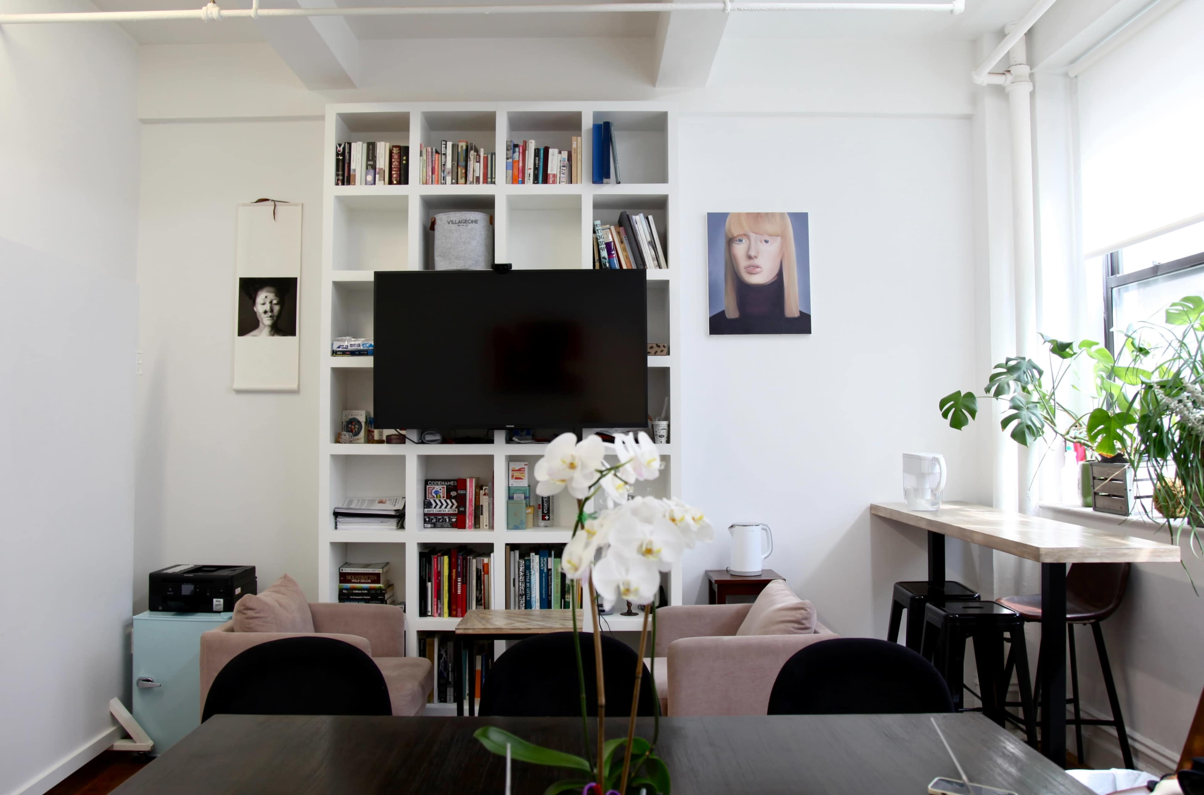 A modern interior features a TV mounted on a white bookshelf filled with books, surrounded by seating and a table with an orchid centerpiece.