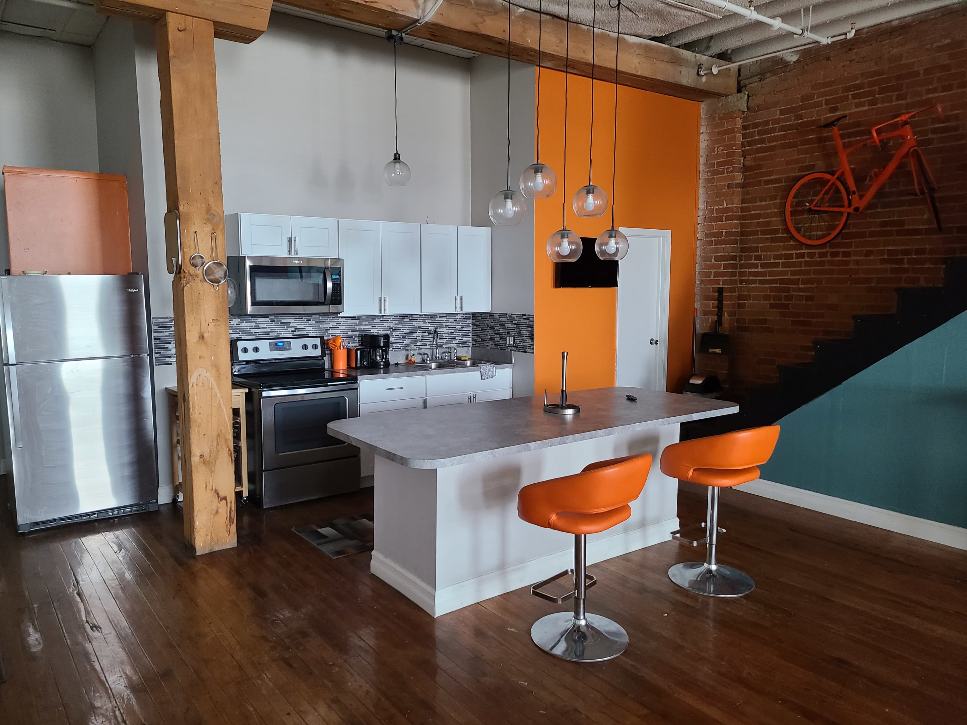 The kitchen features modern appliances, a centrally located island with two orange stools, and a combination of exposed brick and neutral-colored walls.