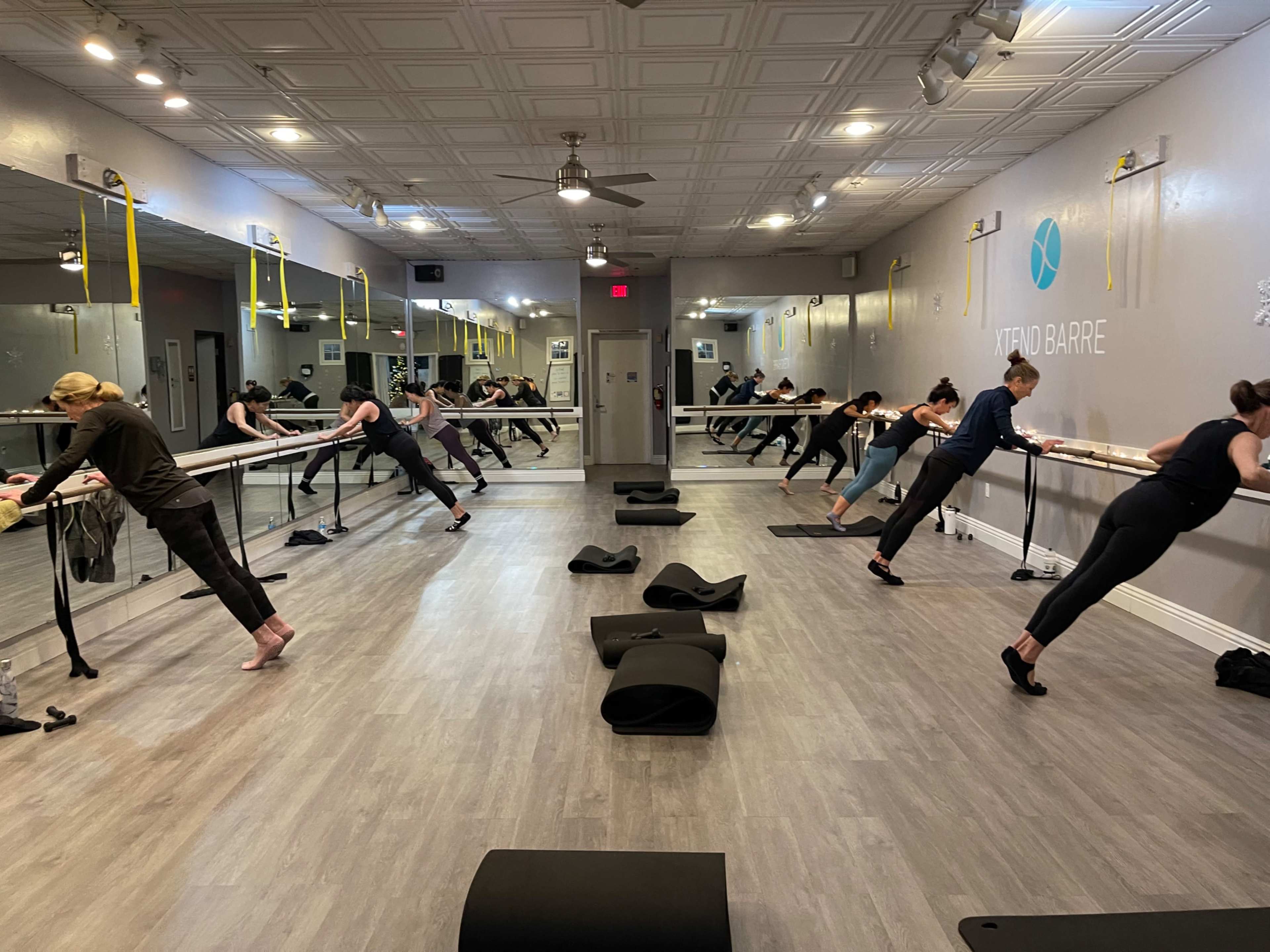 A group of individuals participates in a barre fitness class in a mirrored studio with exercise mats on the floor.