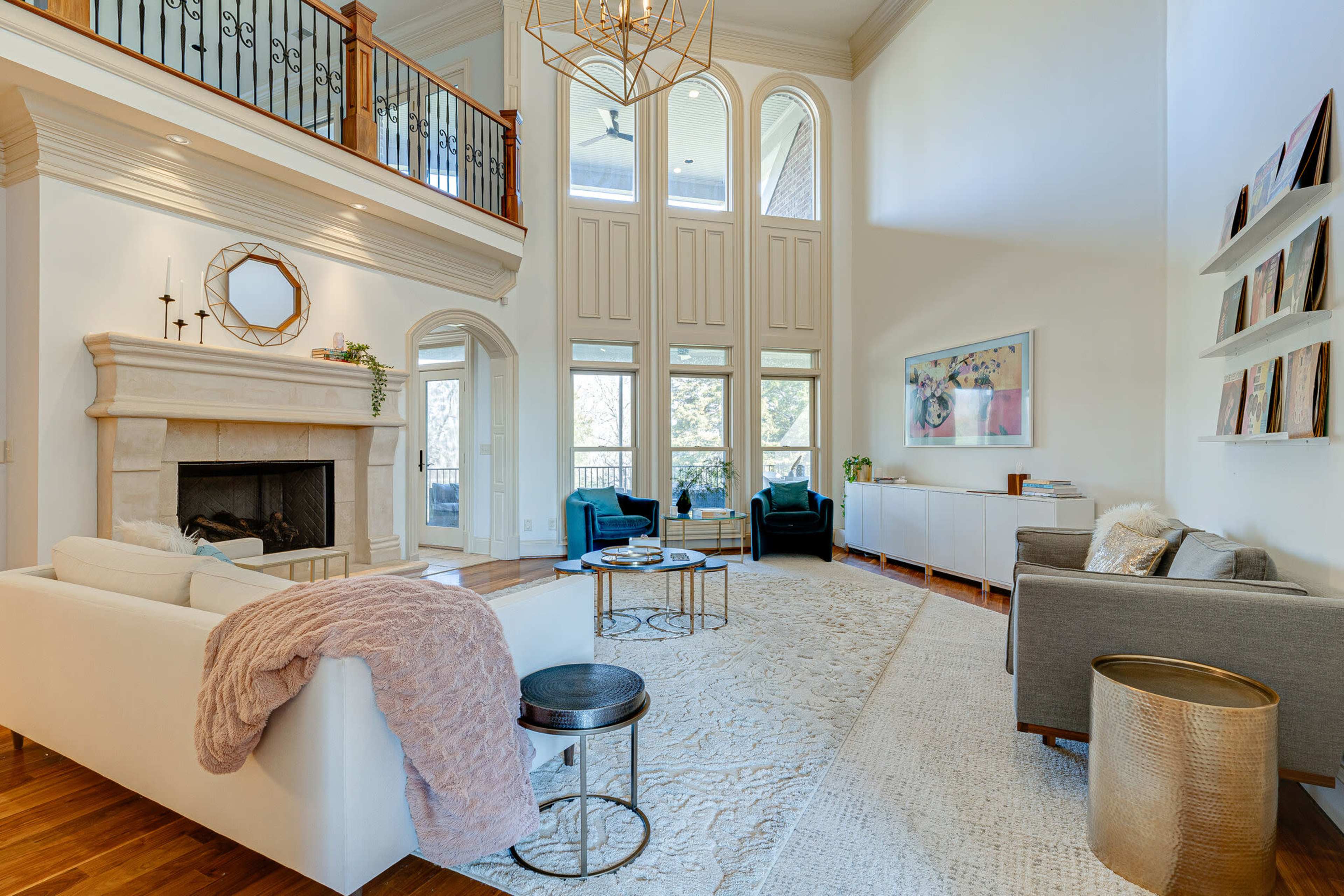 The image shows a spacious living room with high ceilings, featuring a stone fireplace, large windows, and modern furniture arranged around a central area rug.