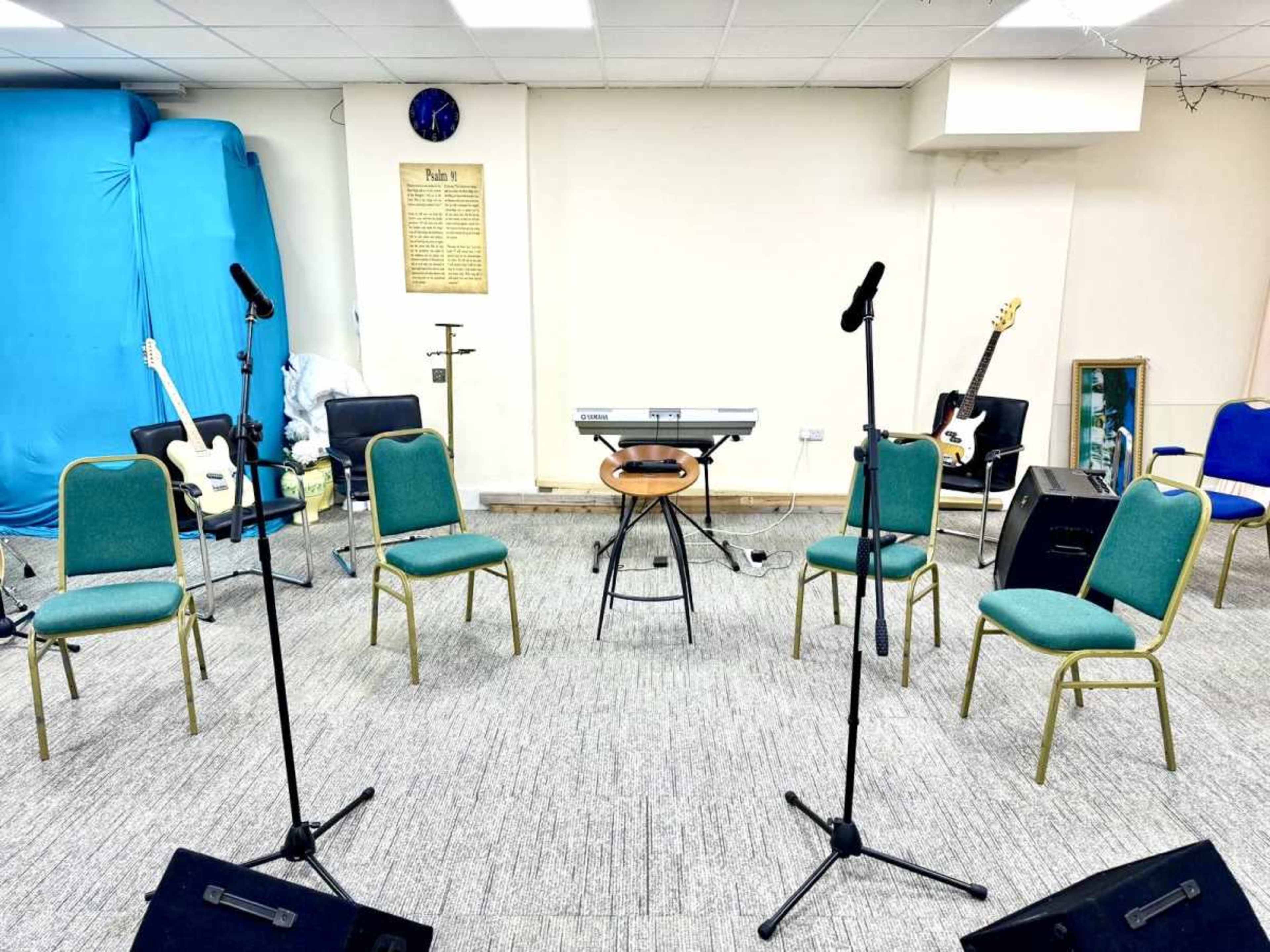 The image shows a room set up for a musical gathering, featuring three microphones, several chairs, a keyboard on a stand, and a guitar against the wall.
