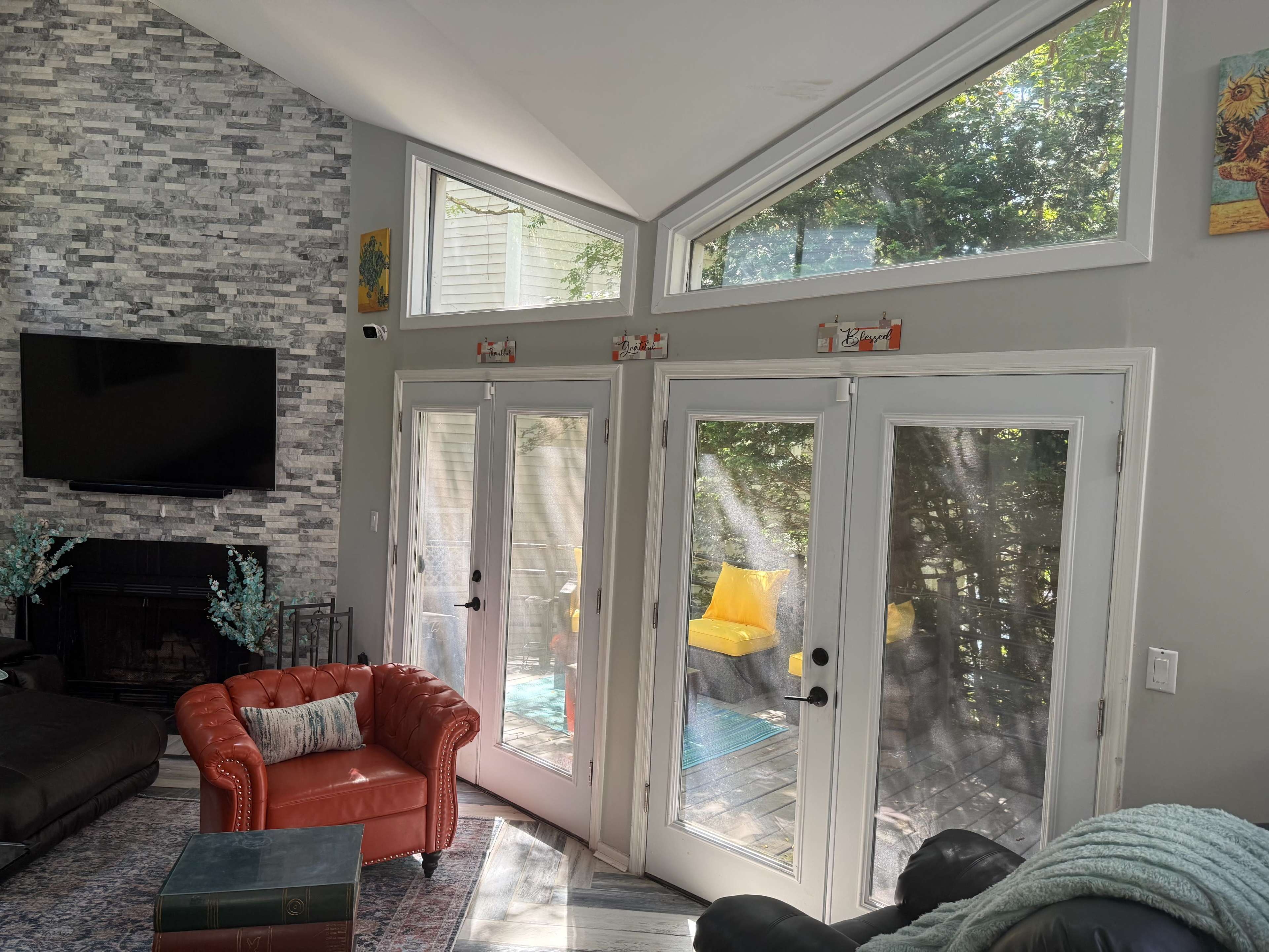 The image shows a modern living room with large windows, a stone fireplace, and a red leather couch facing glass doors that open to an outdoor deck.
