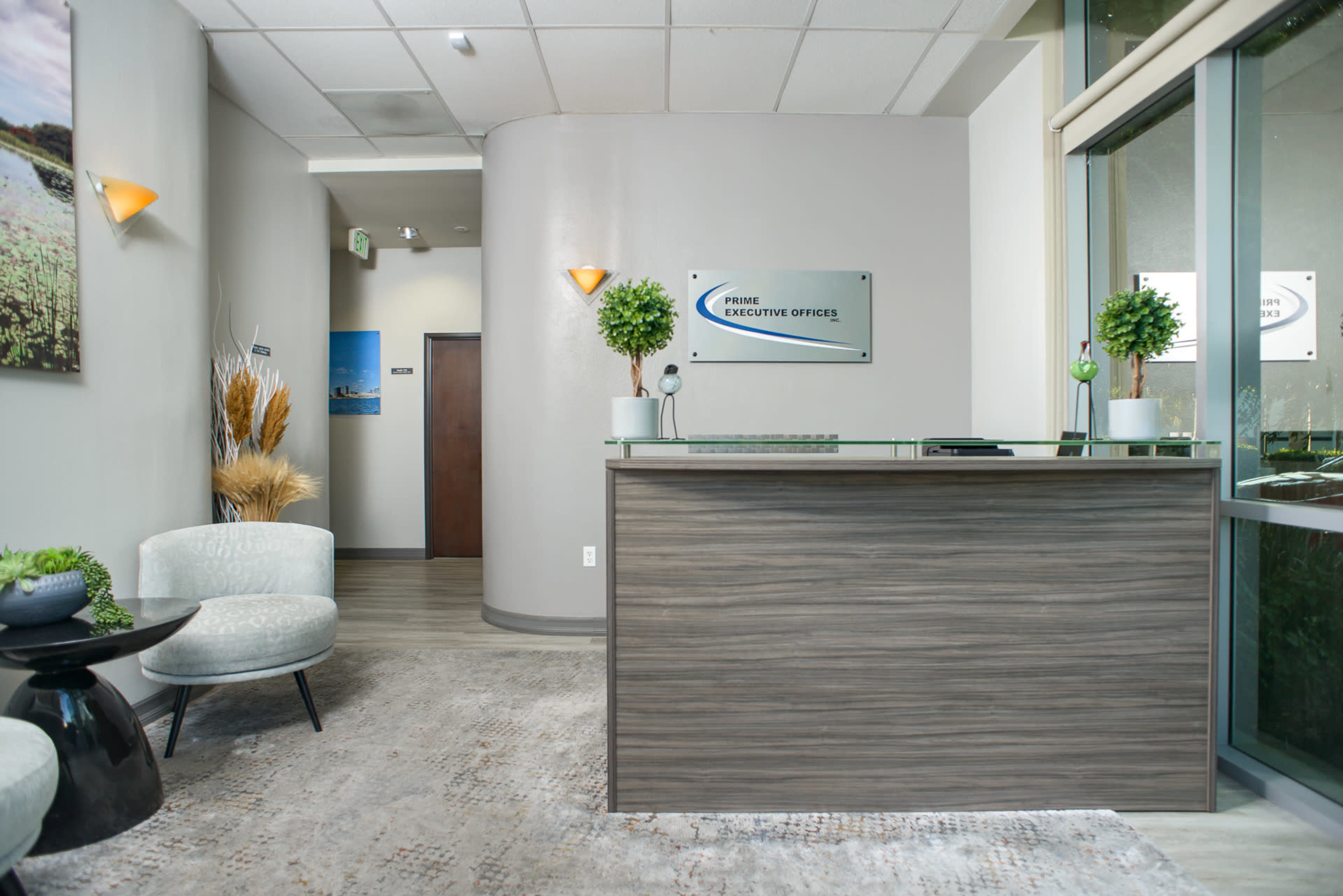 The image shows a modern reception area with a wooden front desk, plants, and seating arrangements.