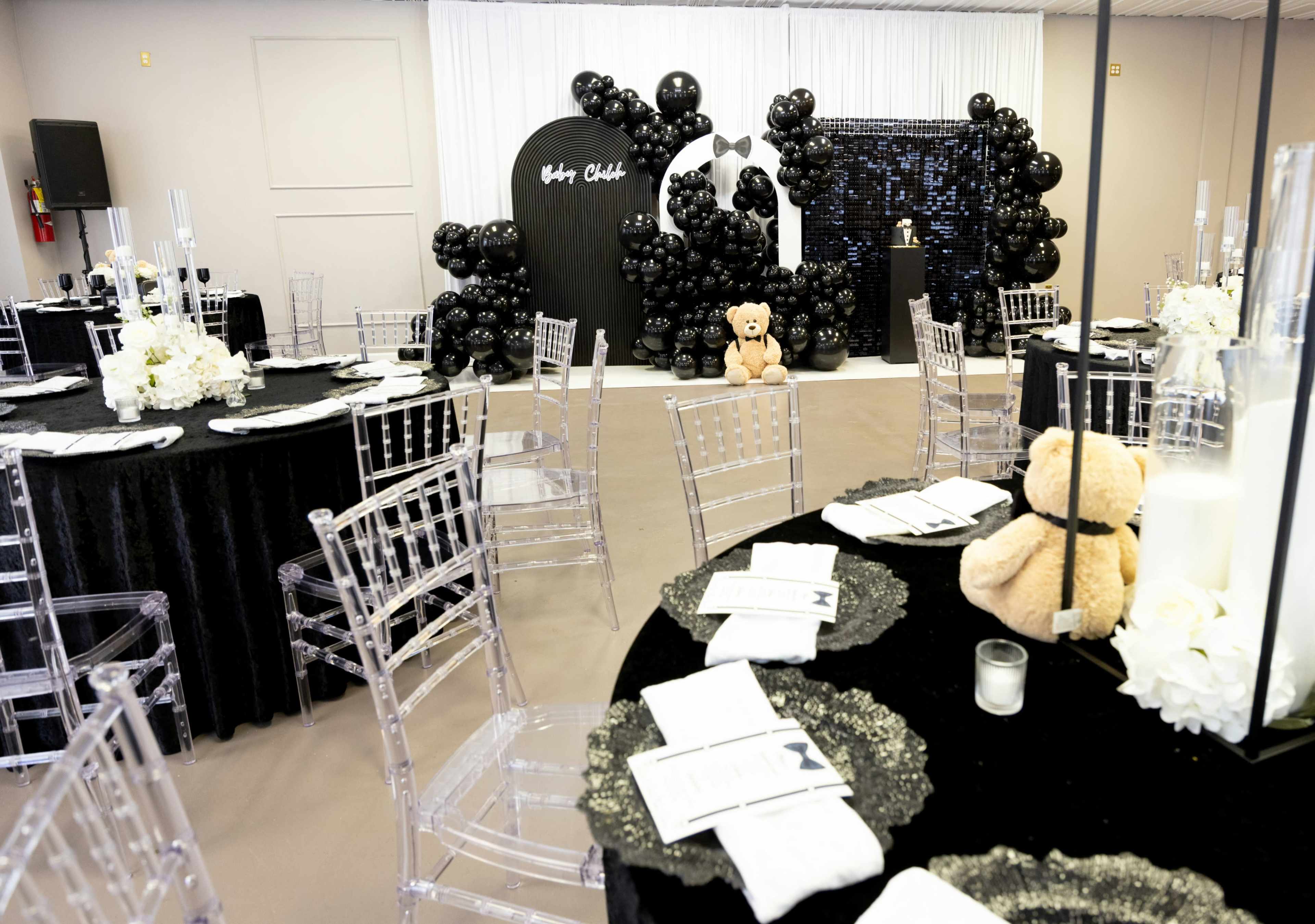 The image shows a banquet room set up for an event with black and white decor, featuring tables with black tablecloths, clear acrylic chairs, and a backdrop adorned with black balloons and a teddy bear.