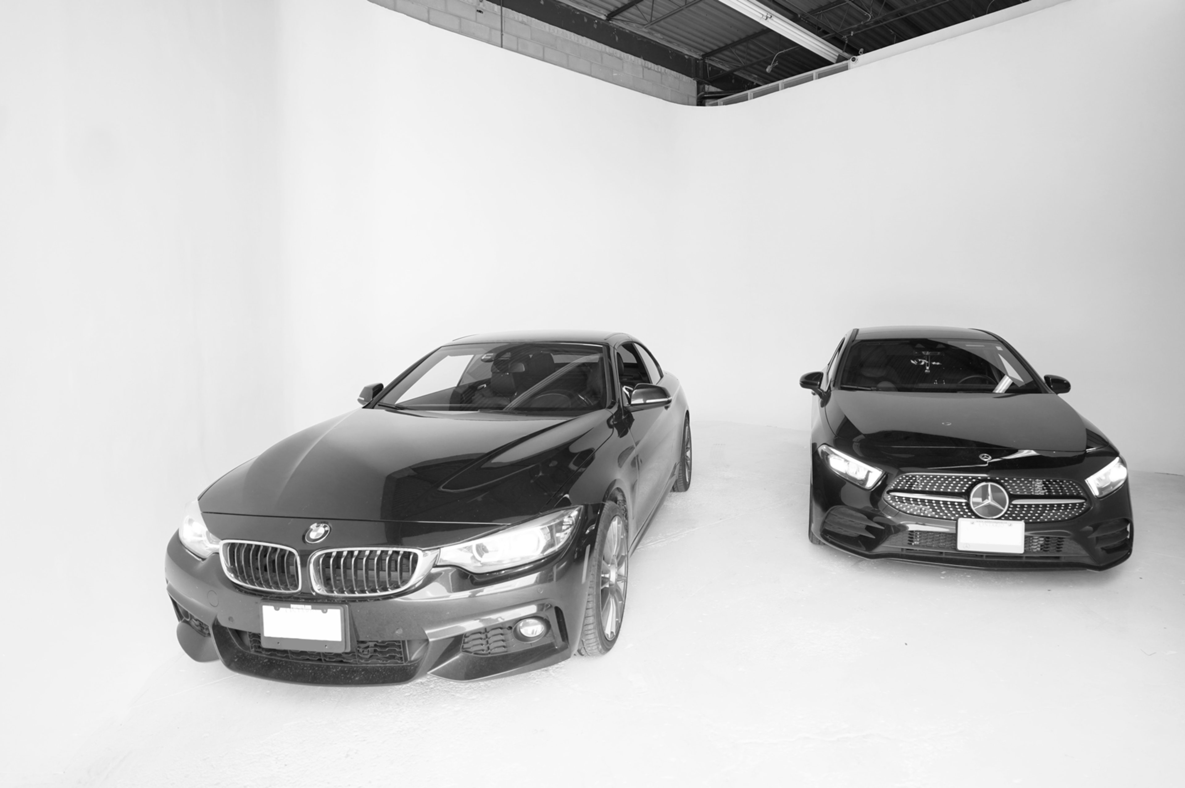 Two luxury cars, a BMW and a Mercedes-Benz, are parked side by side in a minimalistic white studio setting.