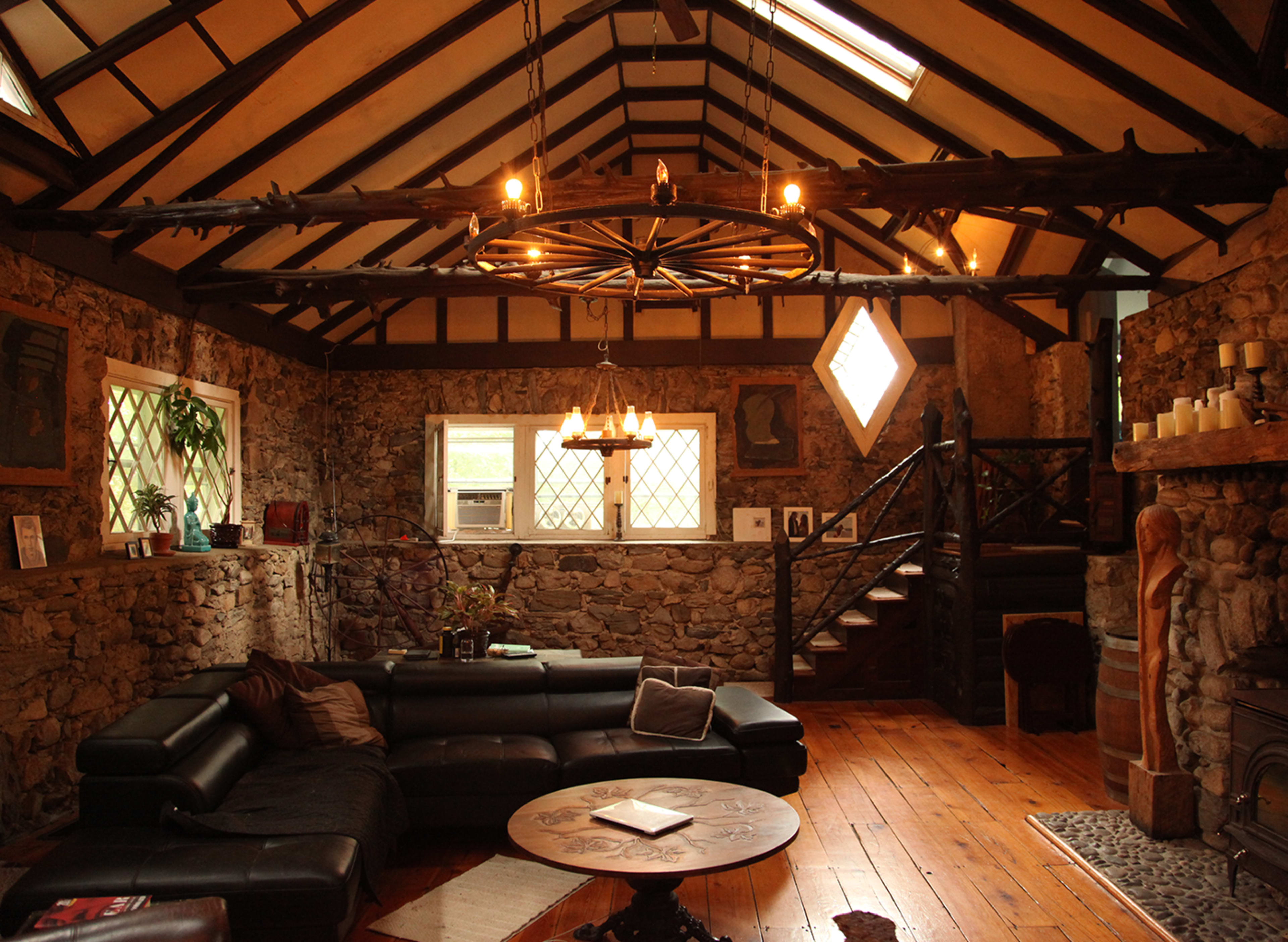 The image shows a rustic living room with stone walls, a wooden ceiling, a sectional sofa, and a staircase leading to an upper level.