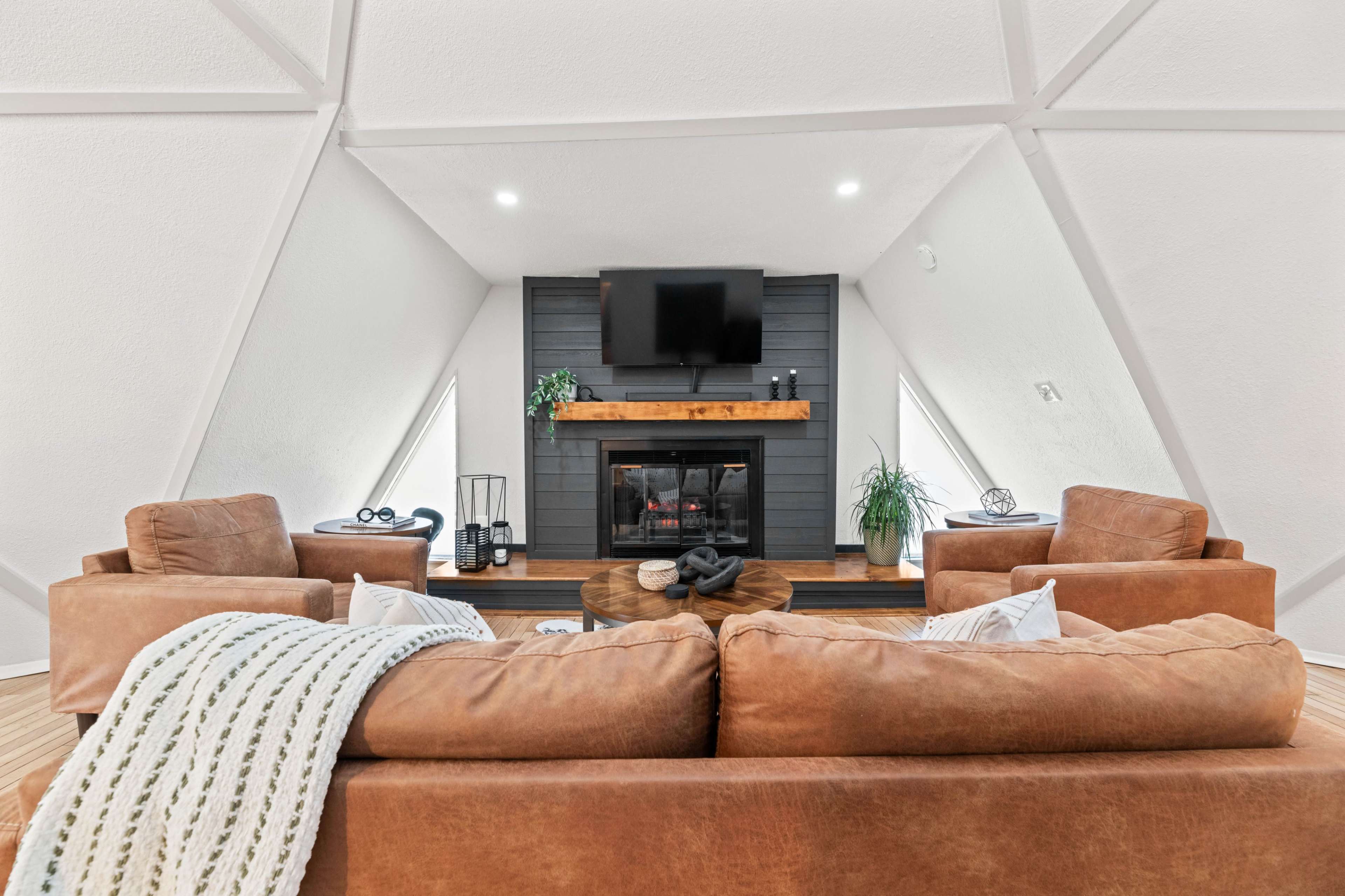 The image shows a cozy living room with a fireplace, a wooden mantel, and two brown leather sofas arranged around a central coffee table, all set beneath a sloped, white ceiling.