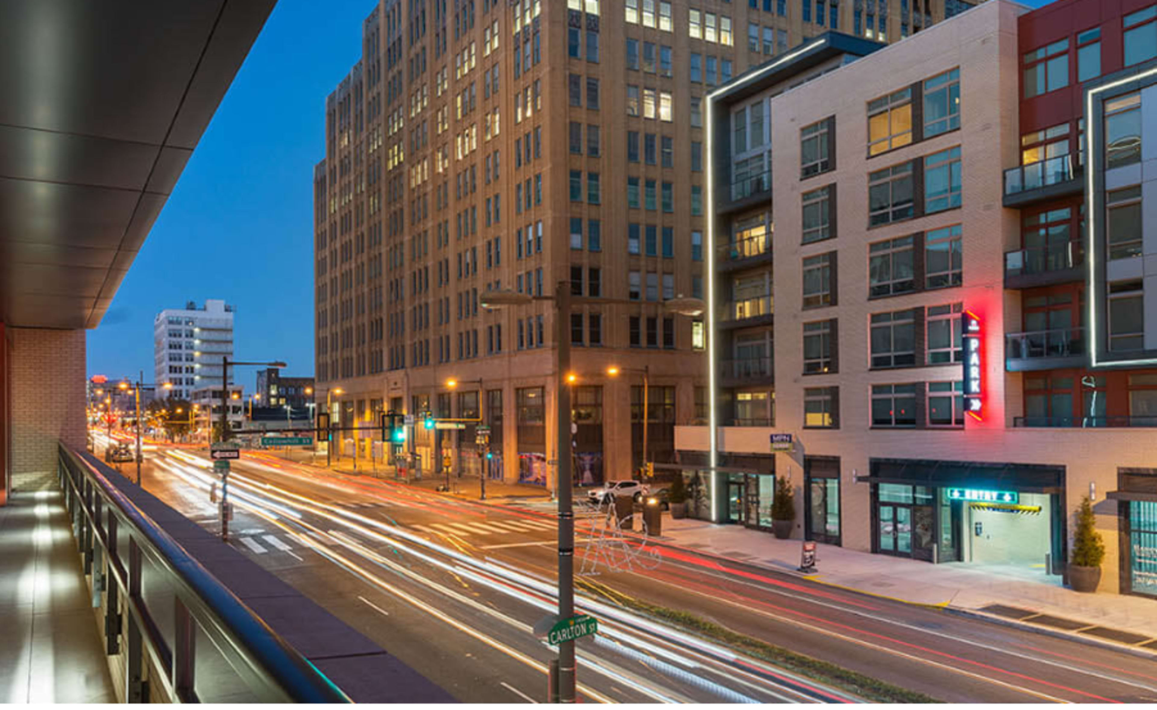 The image shows a city street at dusk, with light trails from moving vehicles and modern buildings lining the road.