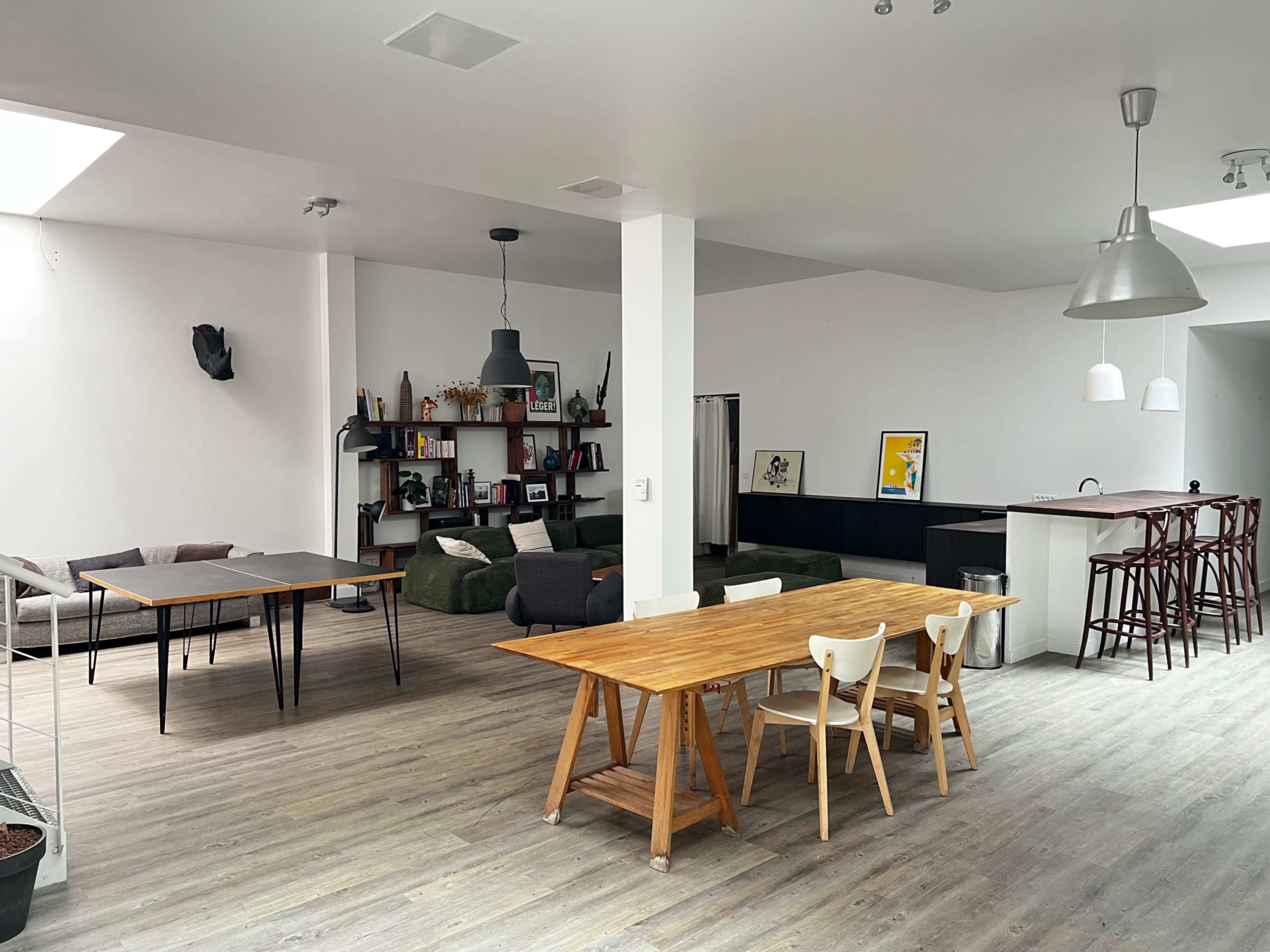 The image shows a spacious, modern open-concept living area featuring a wooden dining table and chairs, a kitchen with bar stools, and a cozy seating arrangement in the background.