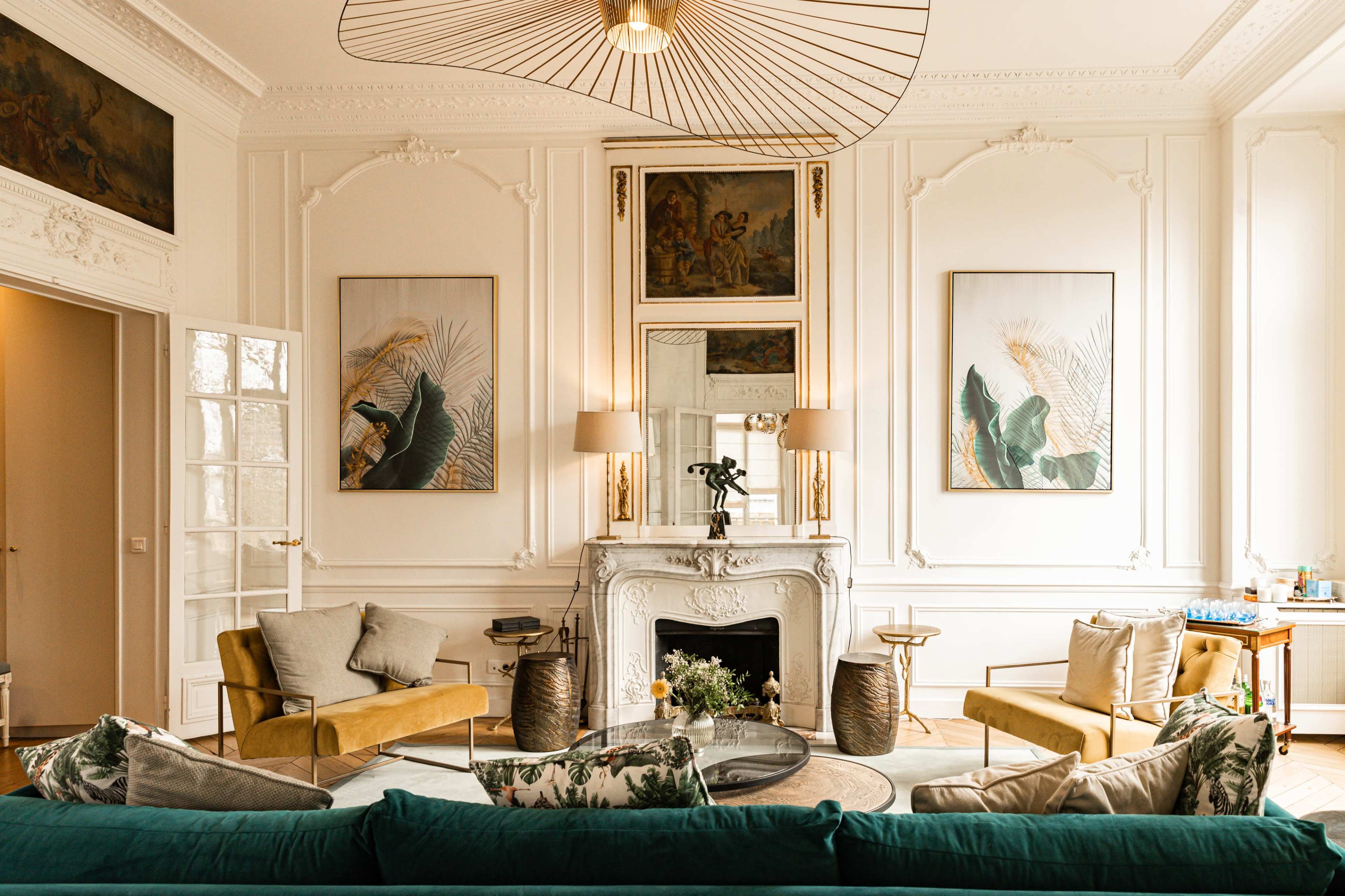 The image shows a stylish living room featuring a white fireplace, two large artworks on the walls, and a mix of upholstered furniture including sofas and armchairs.