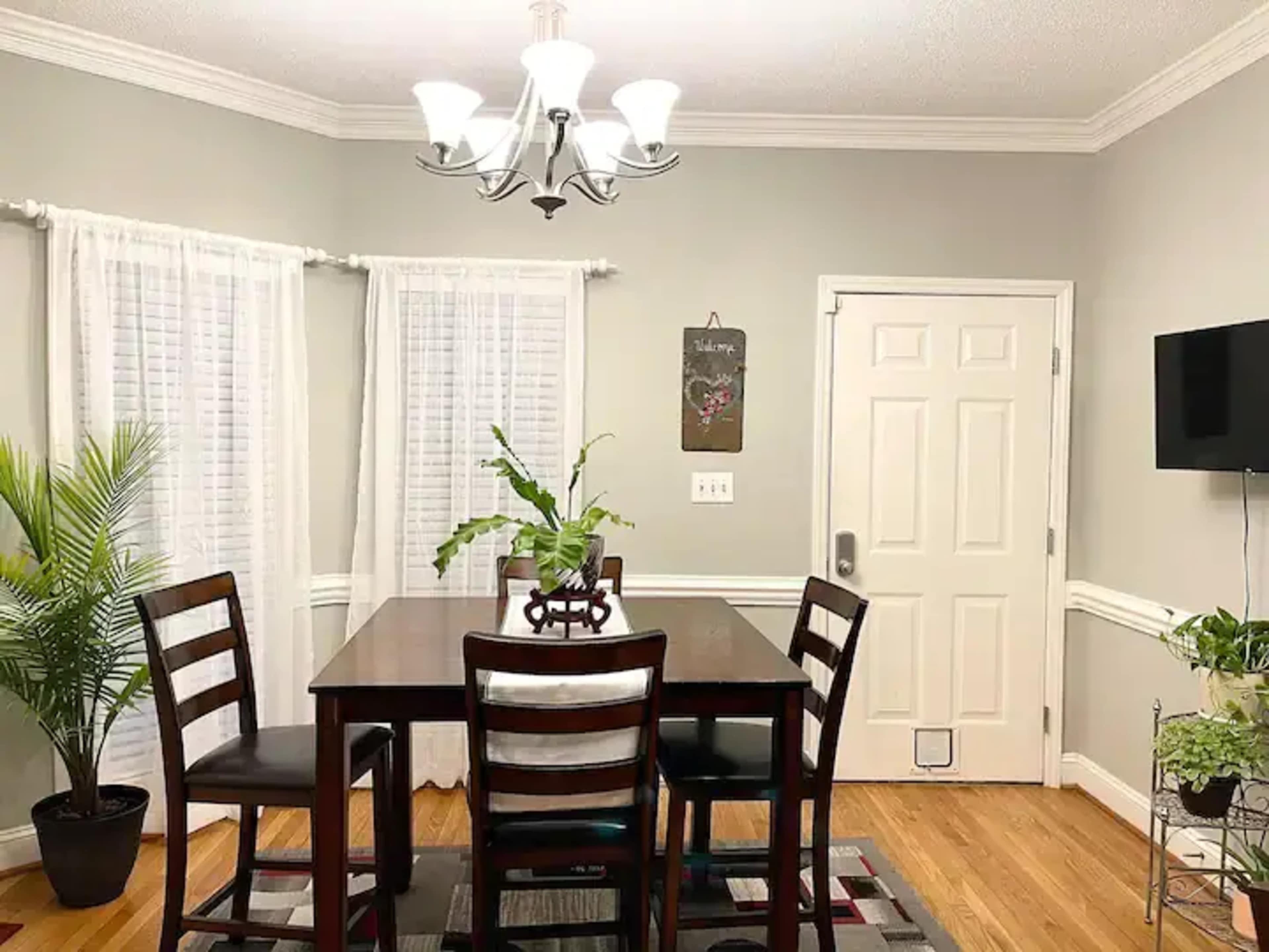 A dining room features a wooden table with six chairs, a chandelier overhead, and a potted plant in the corner.