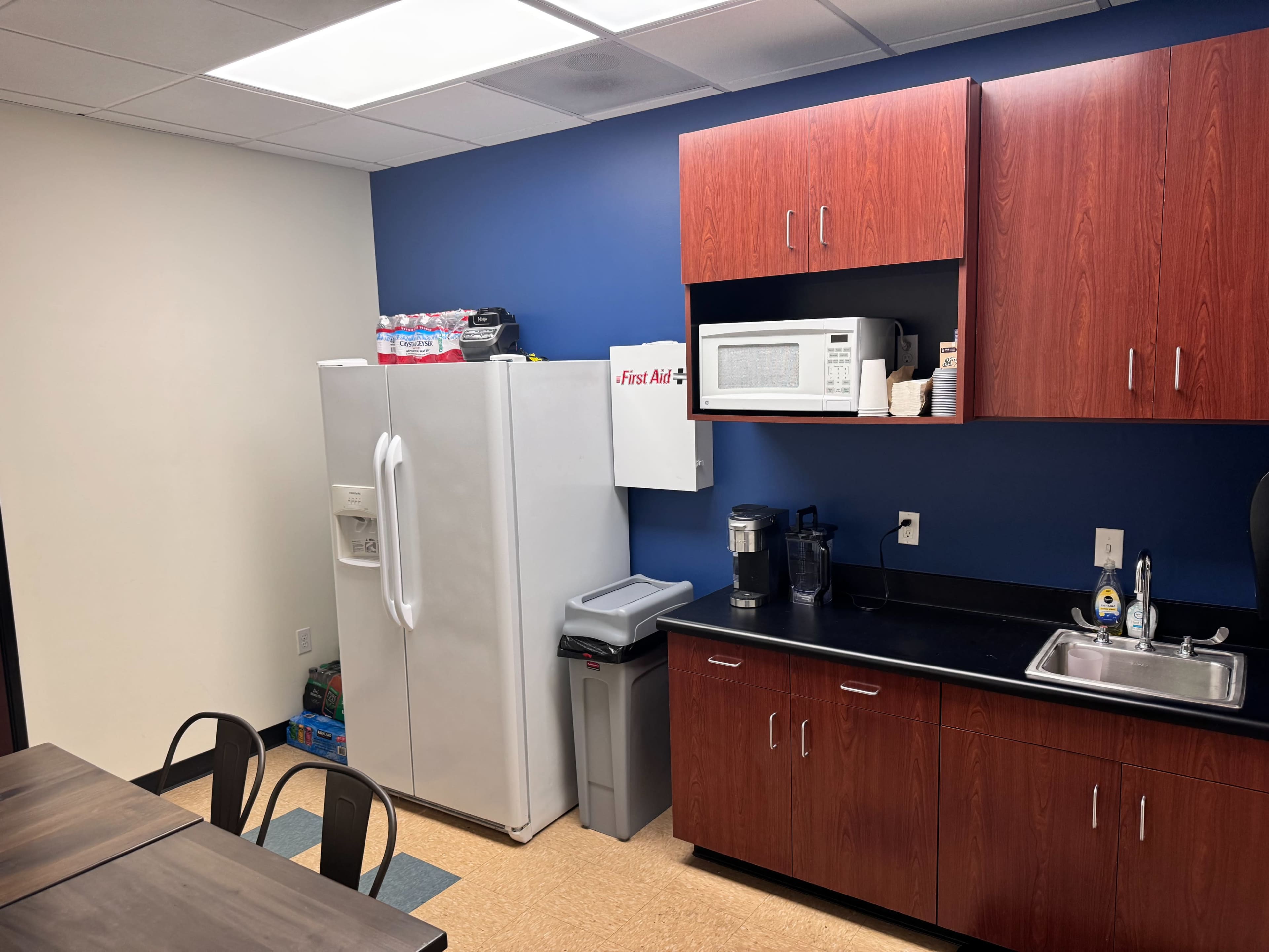 A kitchenette featuring a white refrigerator, a microwave, a sink, and wooden cabinets against a blue wall, with a table and chairs nearby.
