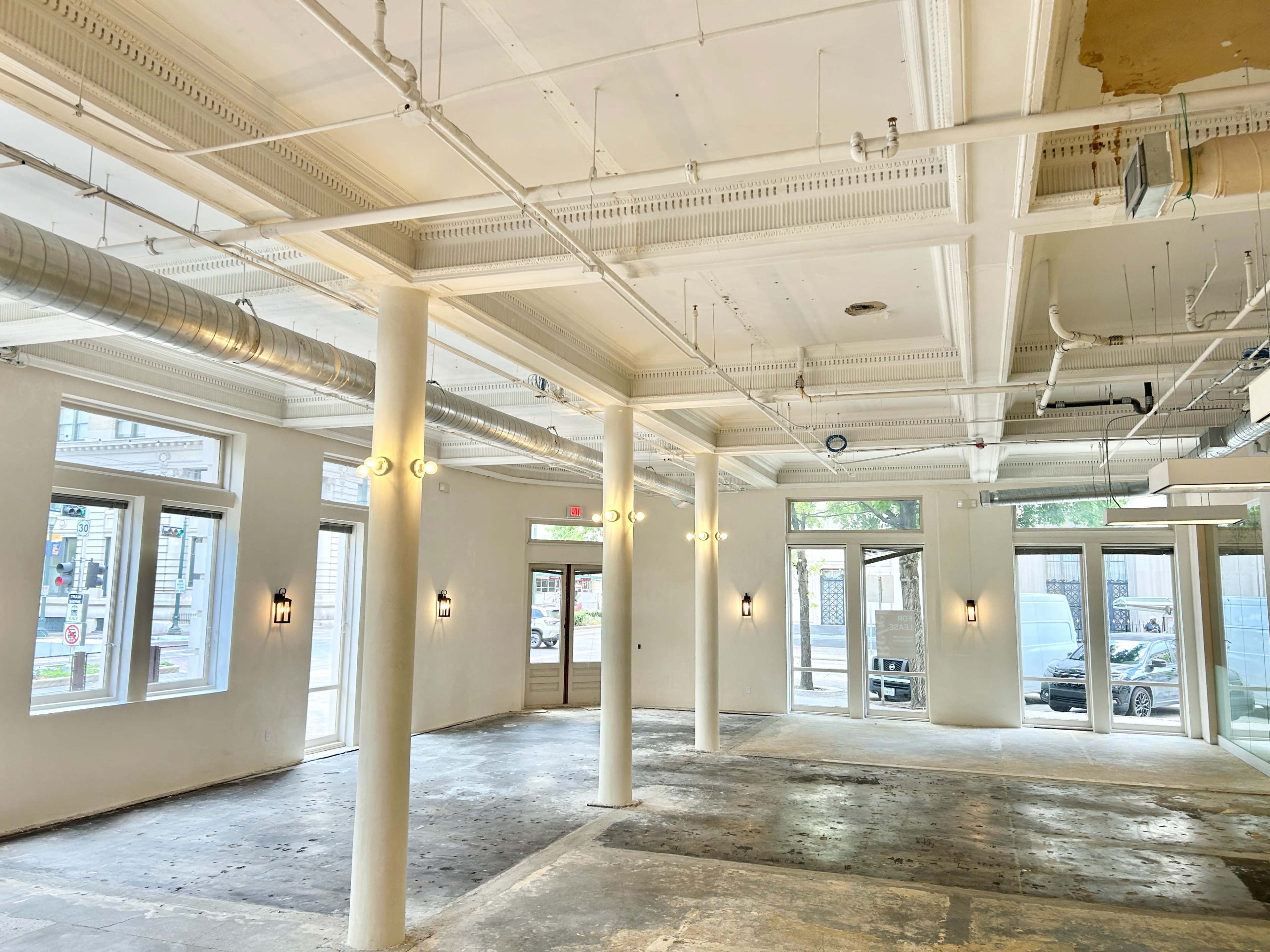The image shows an empty, well-lit commercial space with high ceilings, featuring white columns and large windows.