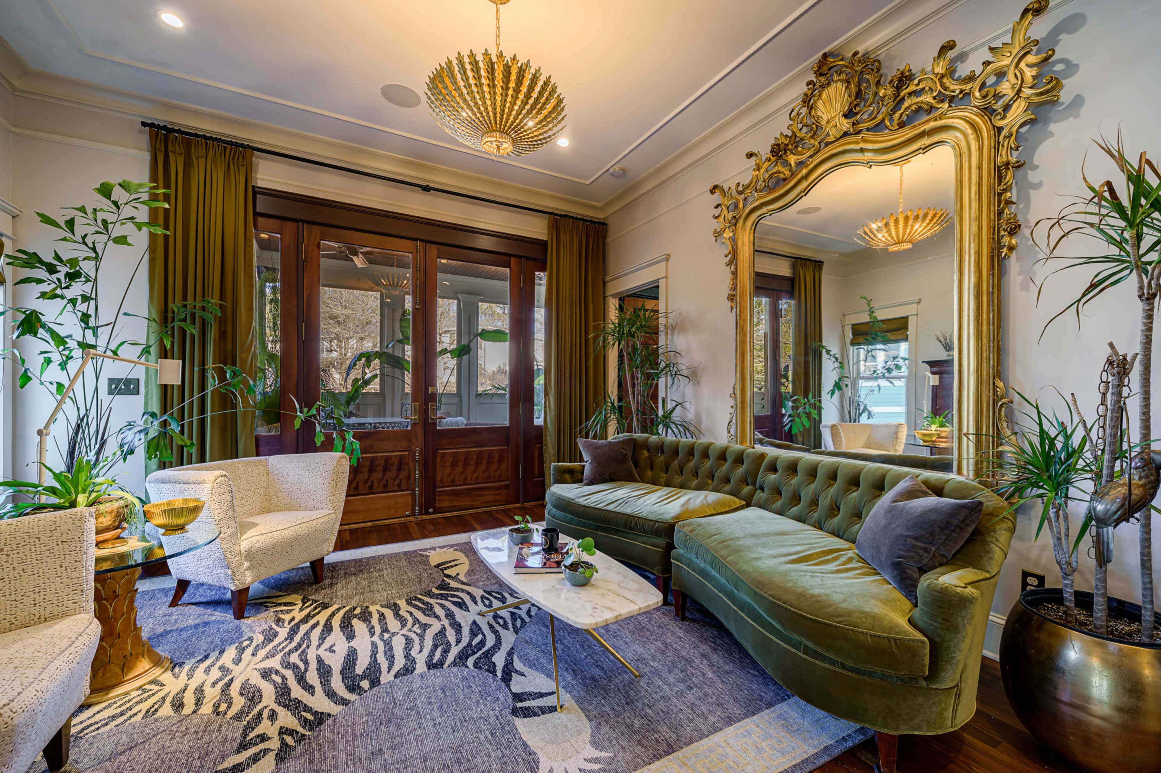 A living room featuring a large green velvet sofa, ornate mirror, potted plants, and a coffee table with a marble top on a patterned rug.
