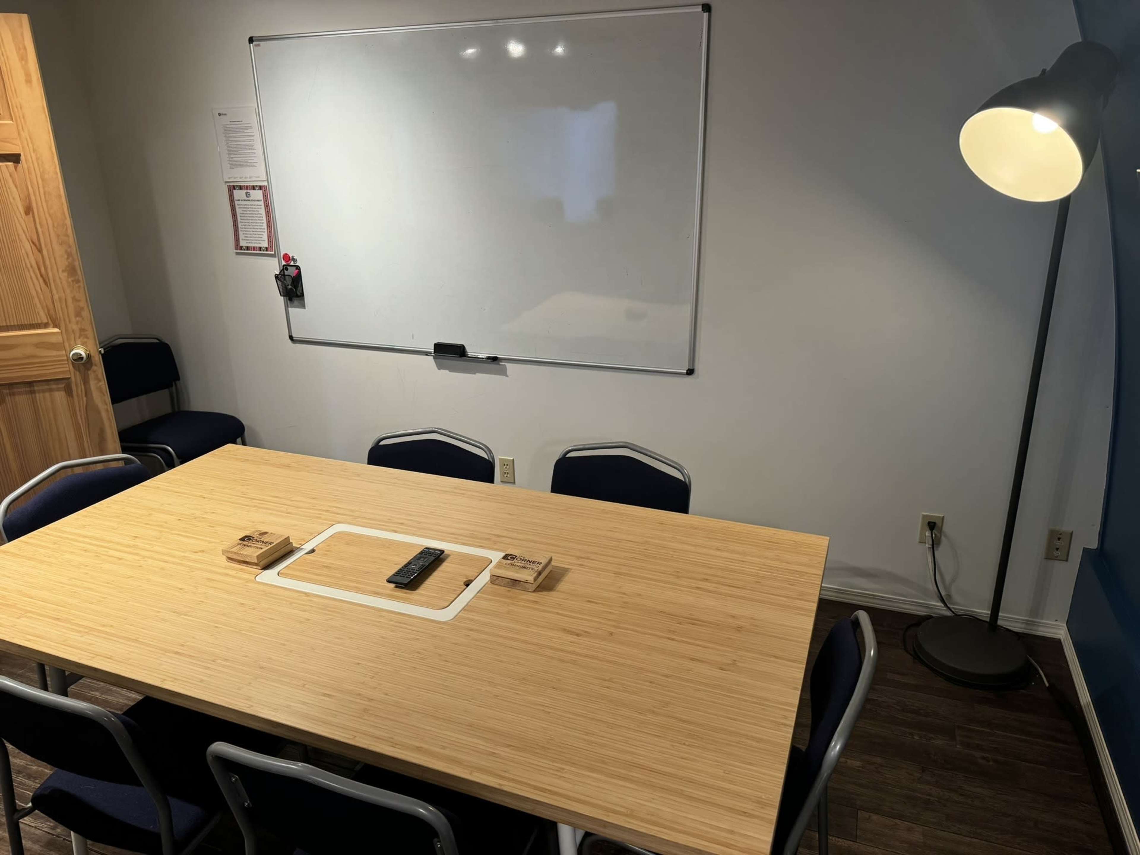 A conference room features a large wooden table, surrounded by blue chairs, with a whiteboard and a lamp in the background.