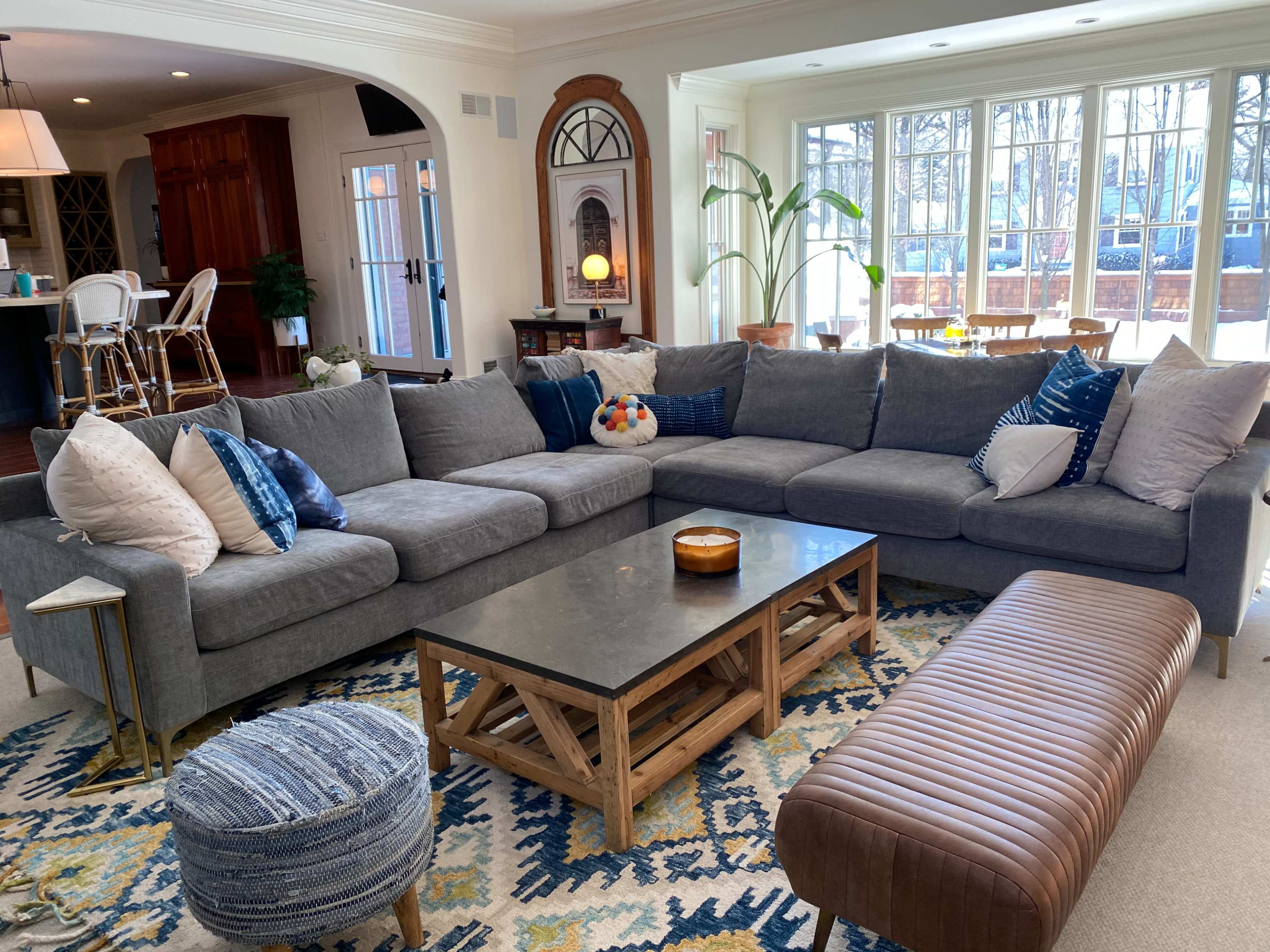 The image shows a modern living room with a large gray sectional sofa, a rectangular coffee table, and a patterned rug, surrounded by bright windows.