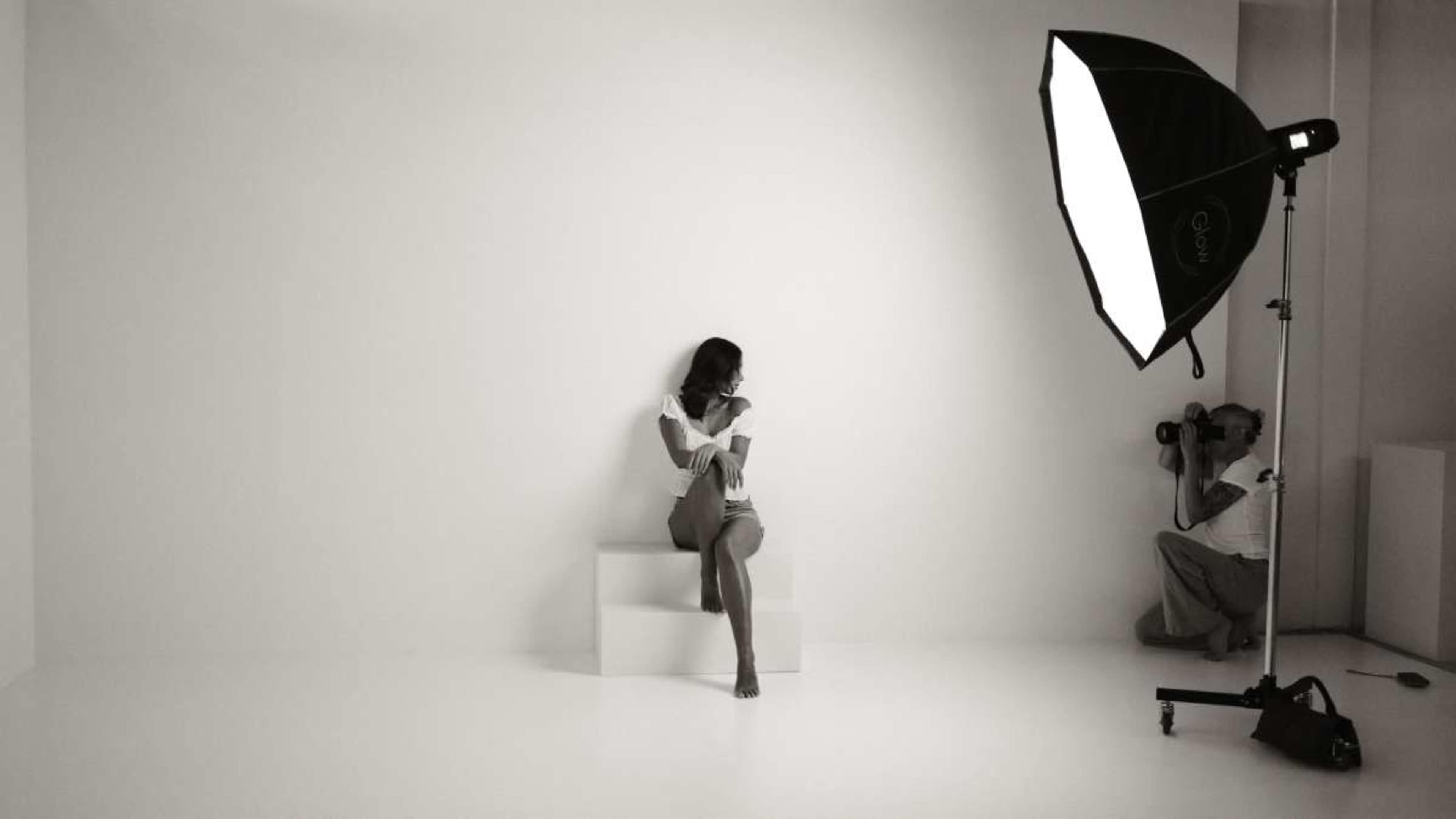 A woman sits on a platform in a minimalistic studio setting while a photographer captures her image.