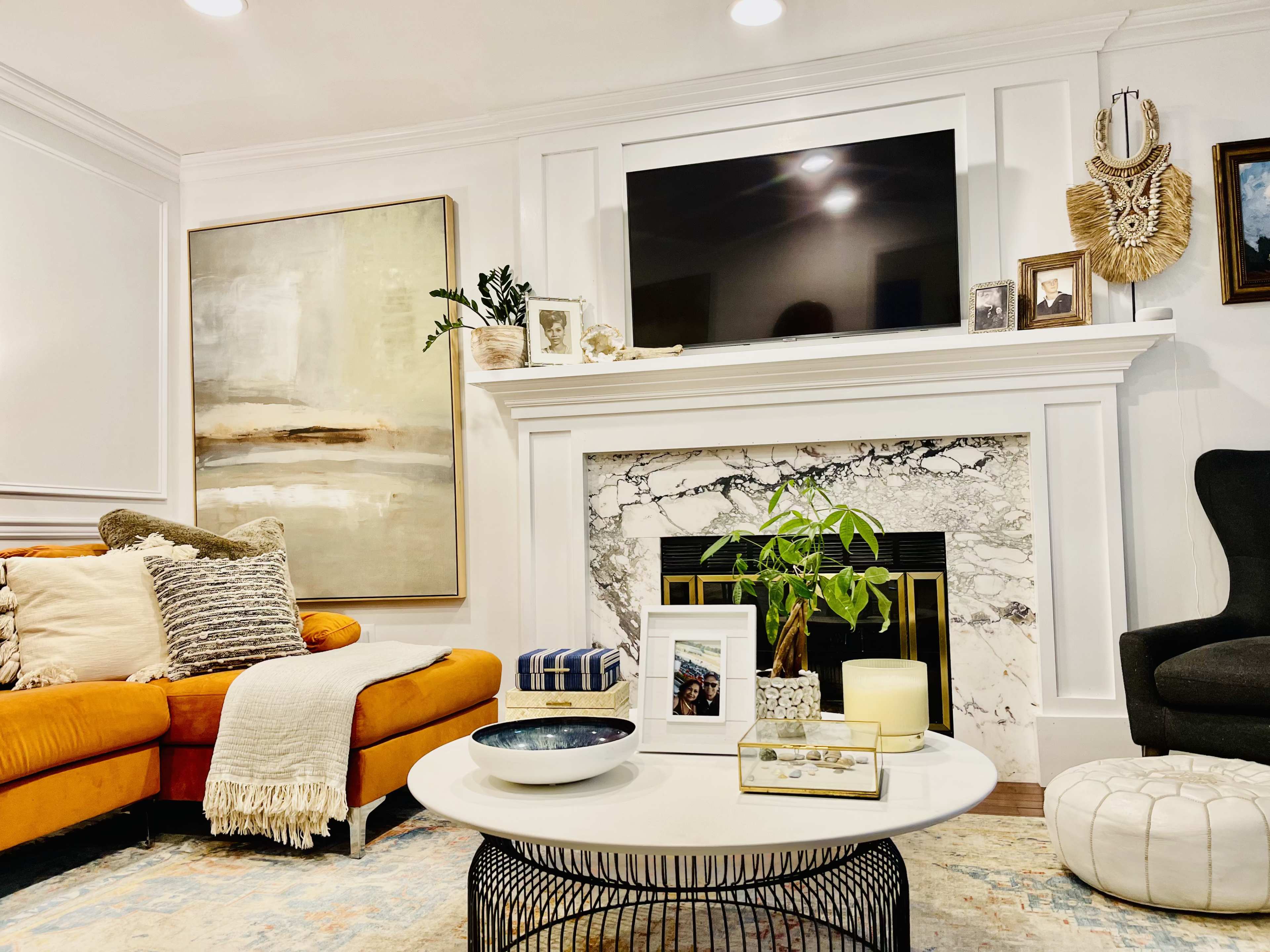The image shows a living room featuring an orange sofa, a round coffee table, and a white marble fireplace with a television mounted above it.