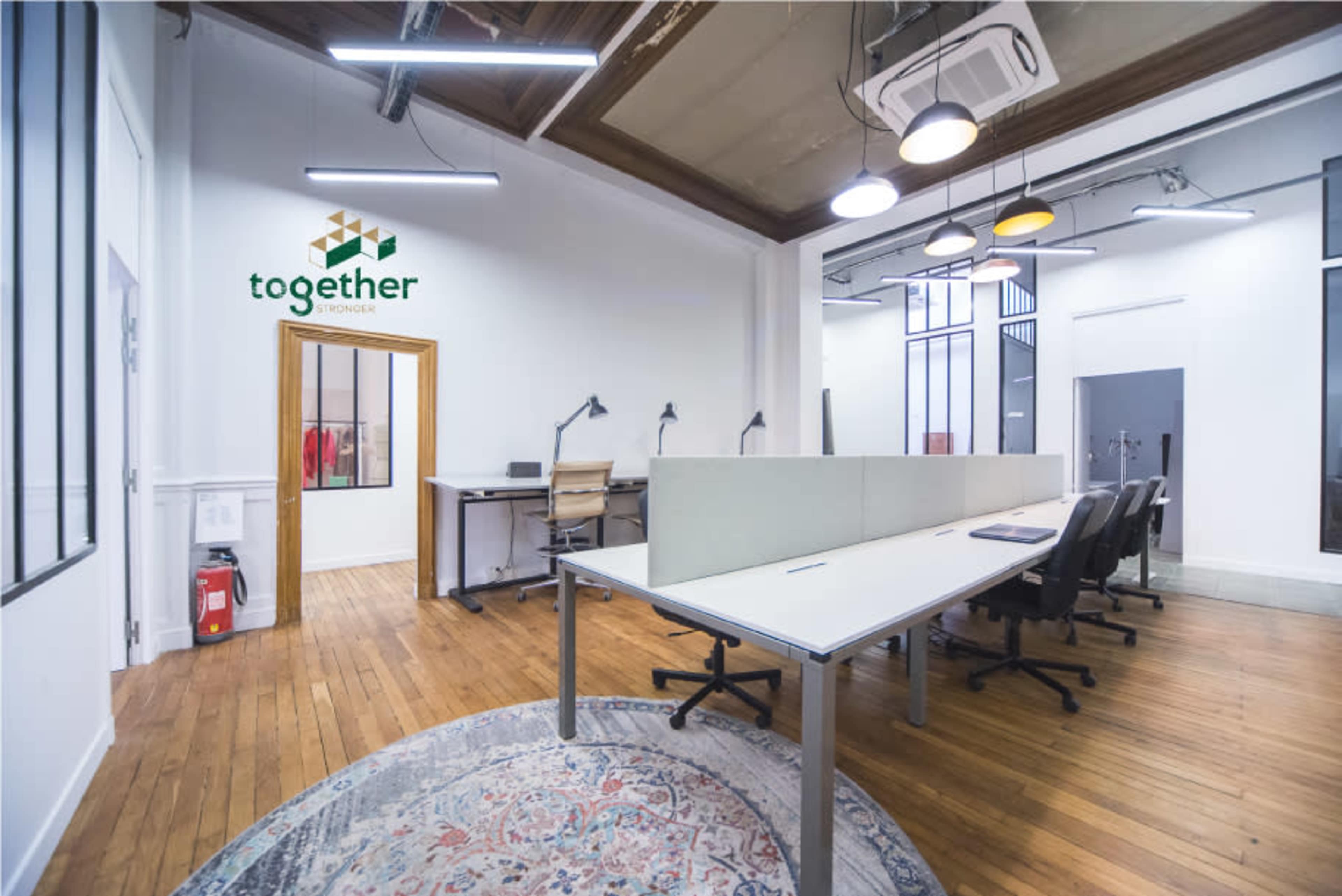 The image shows a modern, well-lit office space with several workstations, wooden flooring, and a decorative rug.