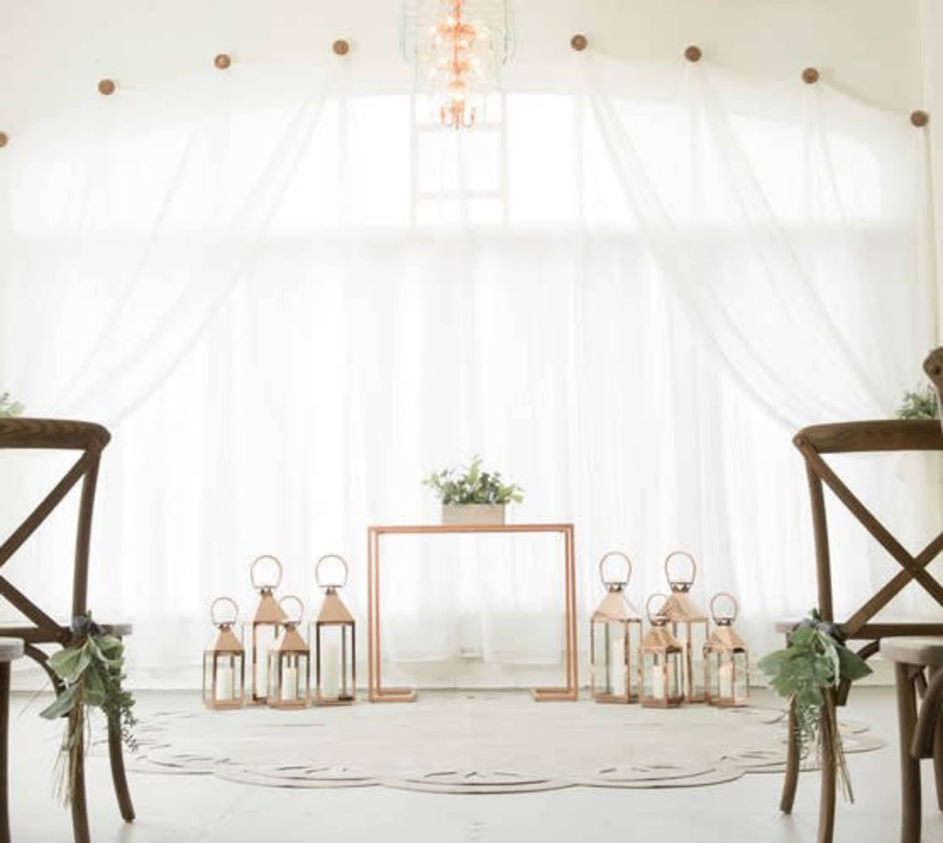 The scene shows a minimalistic wedding setup with wooden chairs facing a small table and decorative lanterns arranged on the floor against a backdrop of draped white fabric.