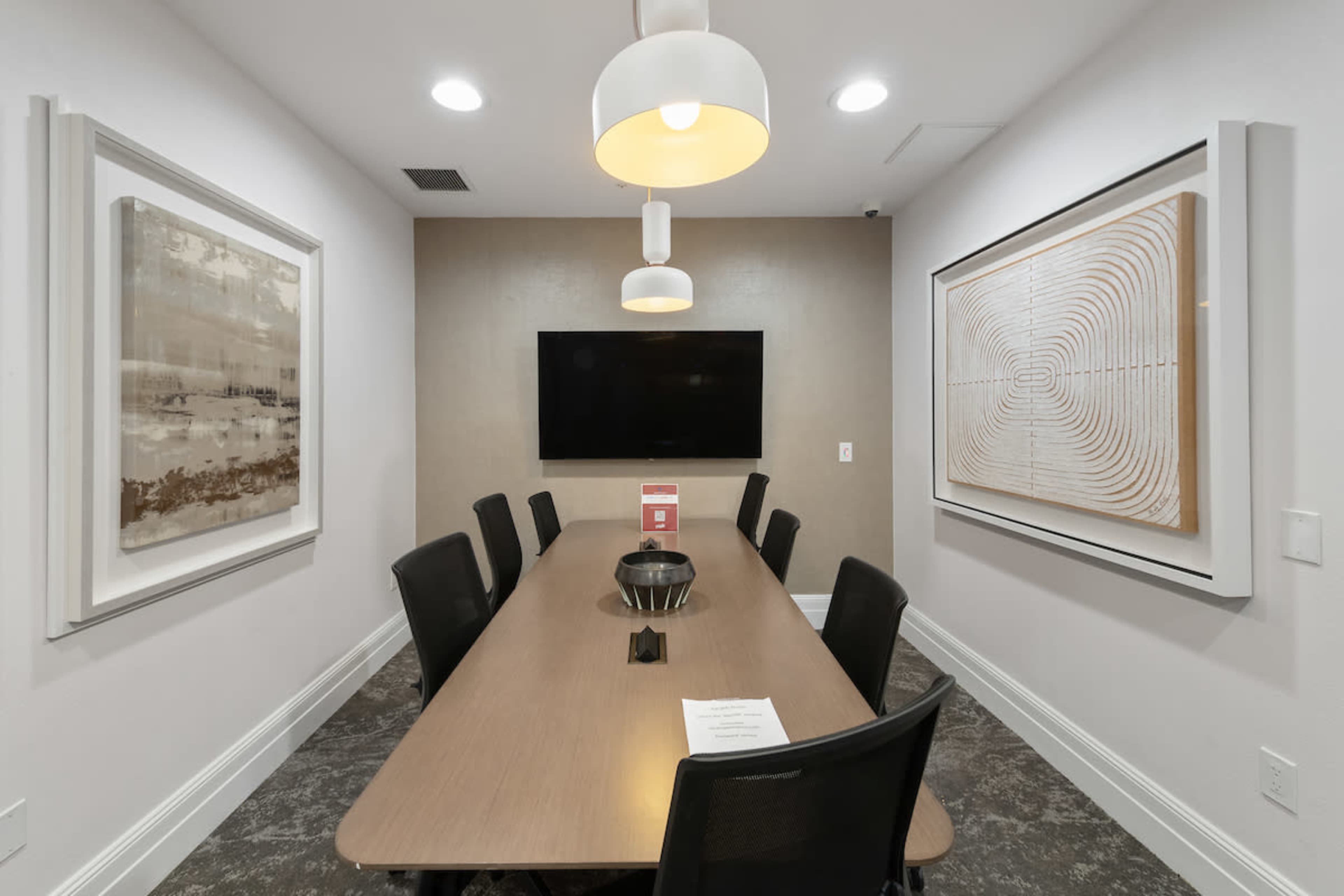 A modern conference room features a long wooden table surrounded by black chairs, with two abstract artworks on the walls and a television mounted at the front.