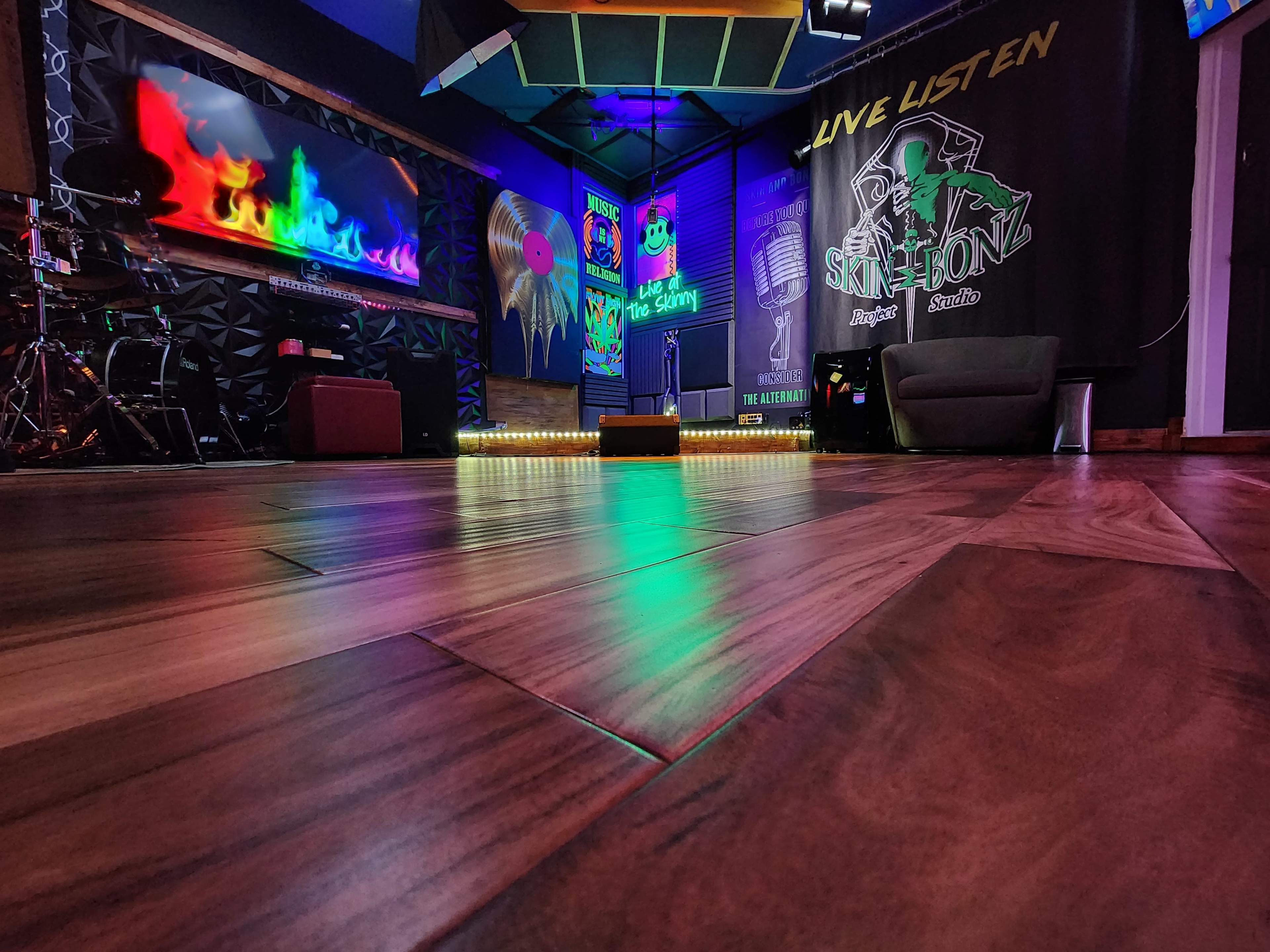 The image shows a recording studio with colorful lighting, wooden flooring, and equipment set up for a live performance.