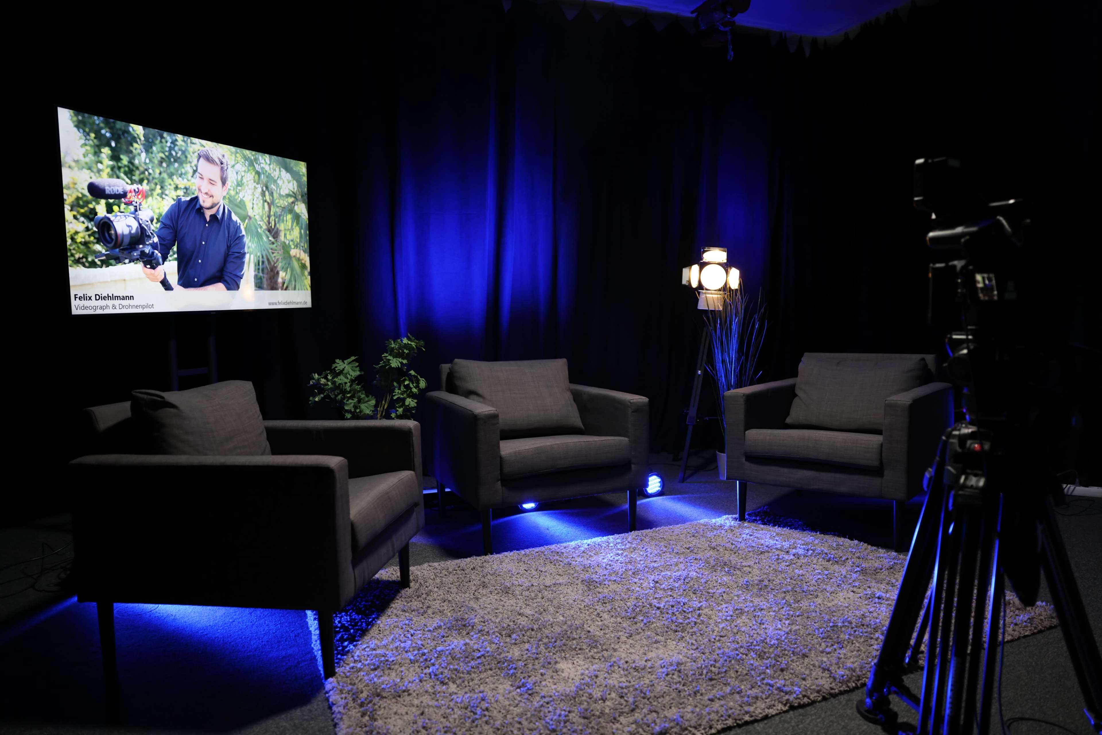 The scene features a modern interview setup with three gray chairs arranged around a rug, a television screen displaying a person with a camera, and a floor lamp providing light in a dark room.