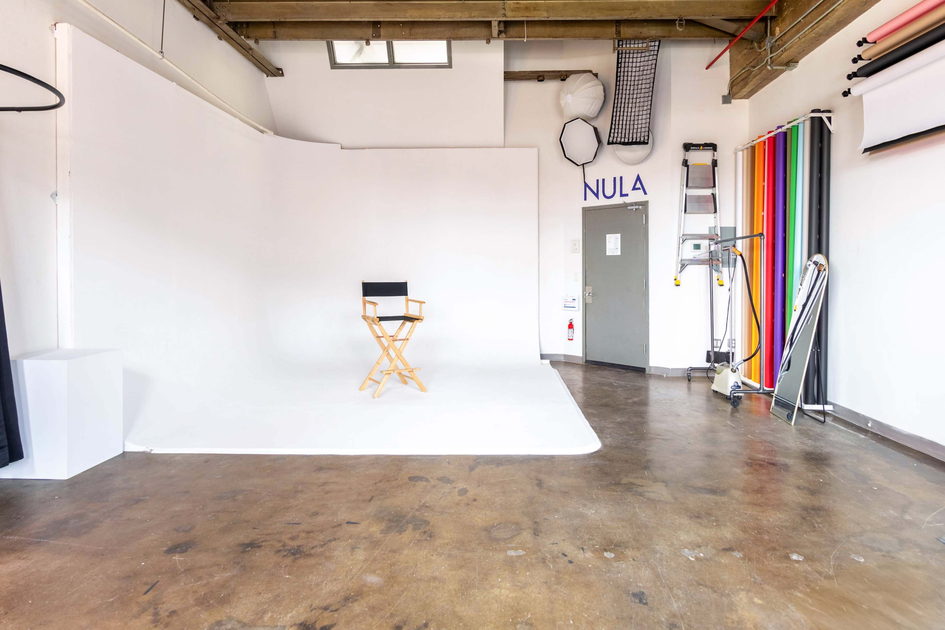 The image shows a photography studio with a white backdrop, a single director's chair, and a wall of colored backdrops on one side.