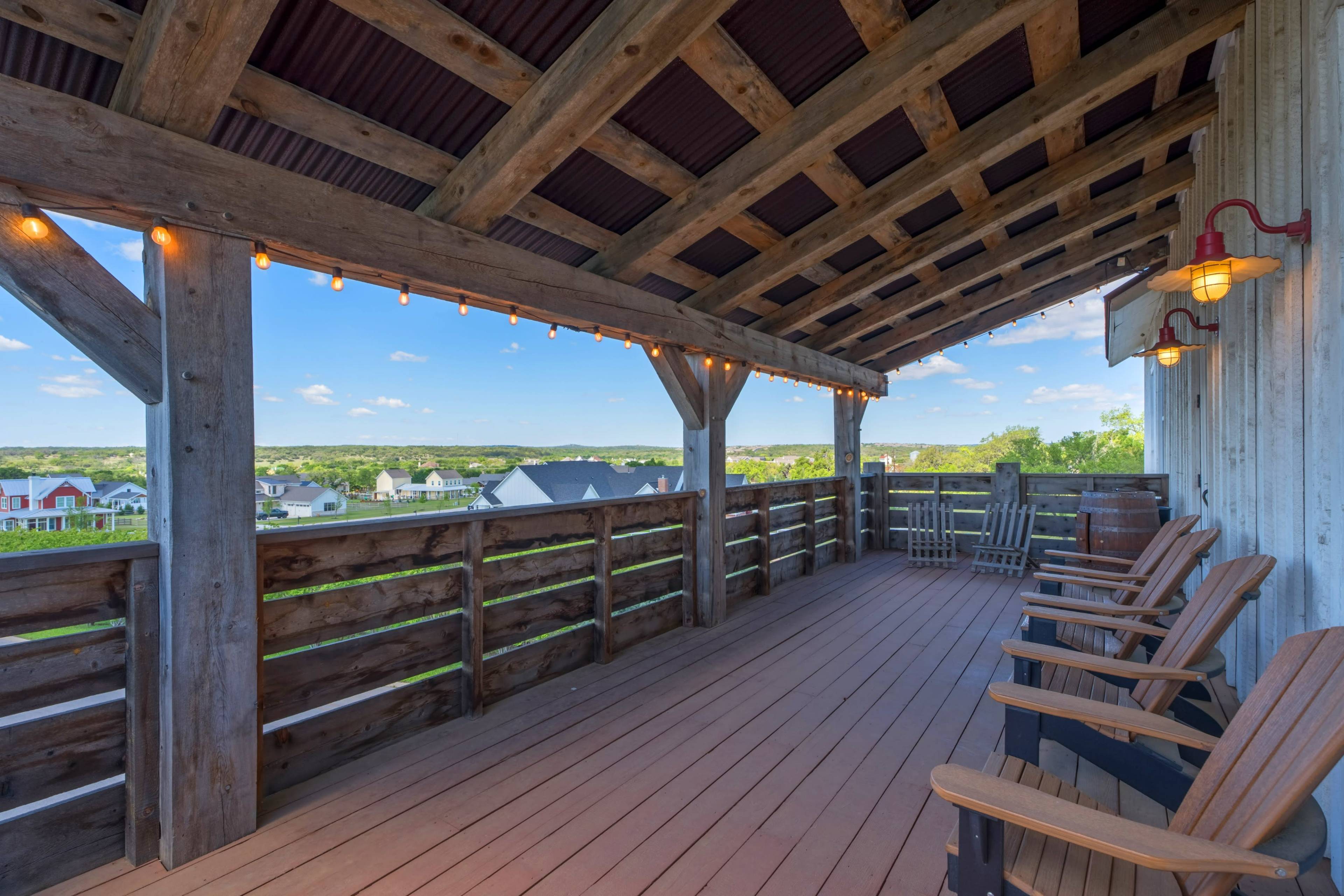 A spacious wooden deck with several rocking chairs, a railing made of wooden beams, and string lights, overlooking a scenic landscape of homes and hills.