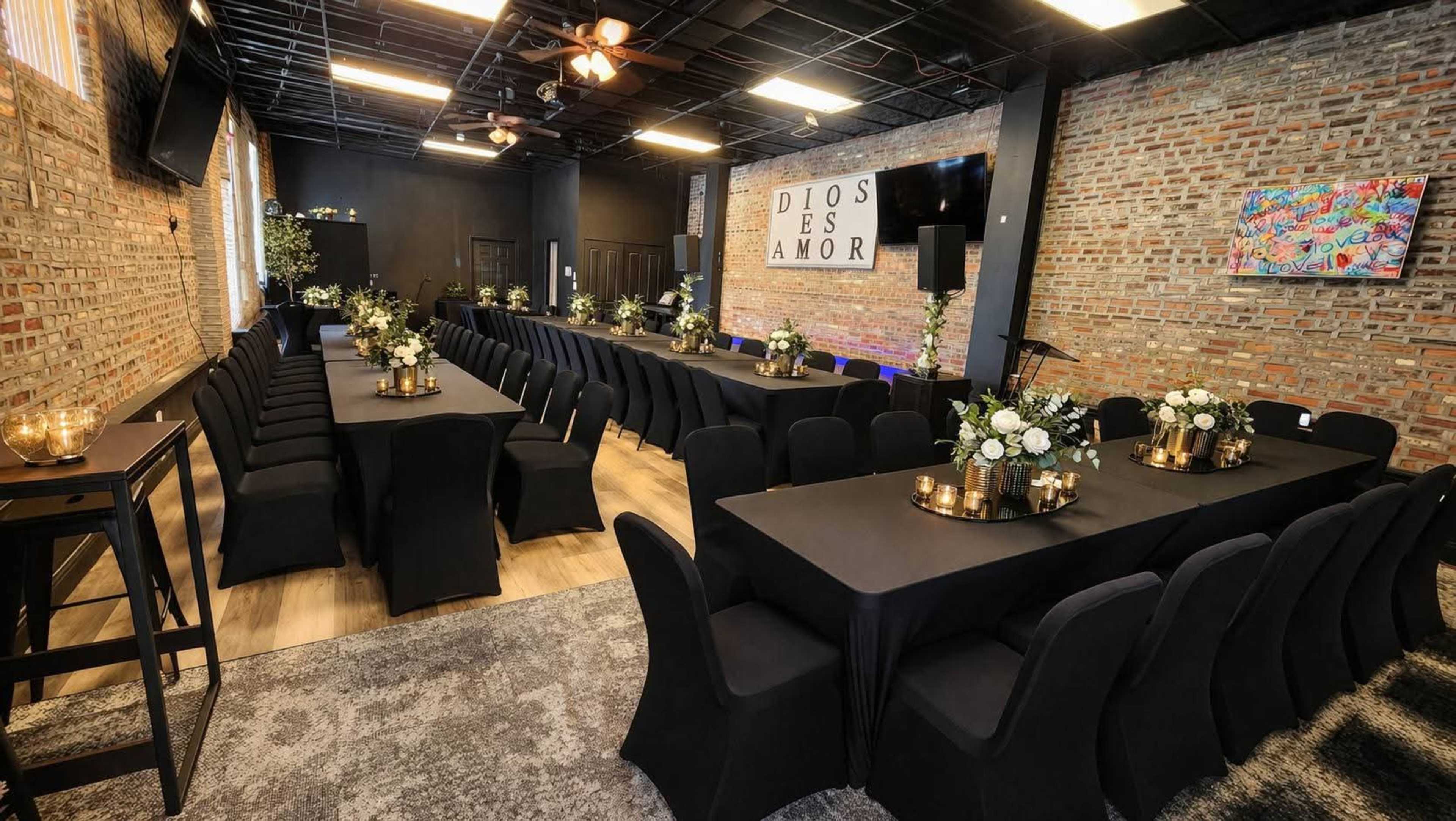 A spacious event room features two long rows of tables set with black tablecloths and floral centerpieces, against a backdrop of exposed brick walls.